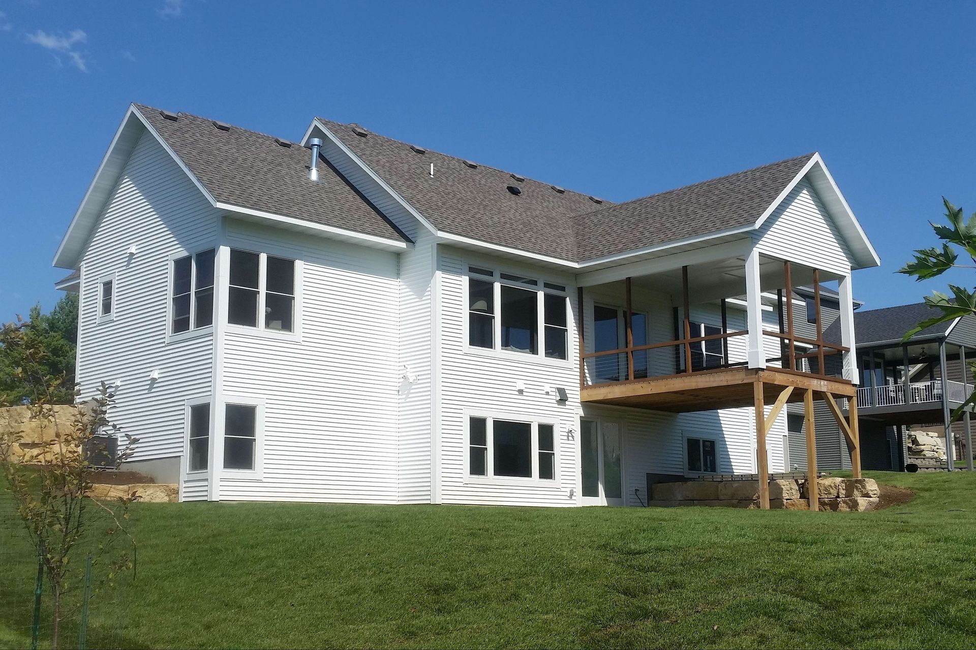 White two-story house with a wooden deck, set on a green grassy hill under a blue sky.