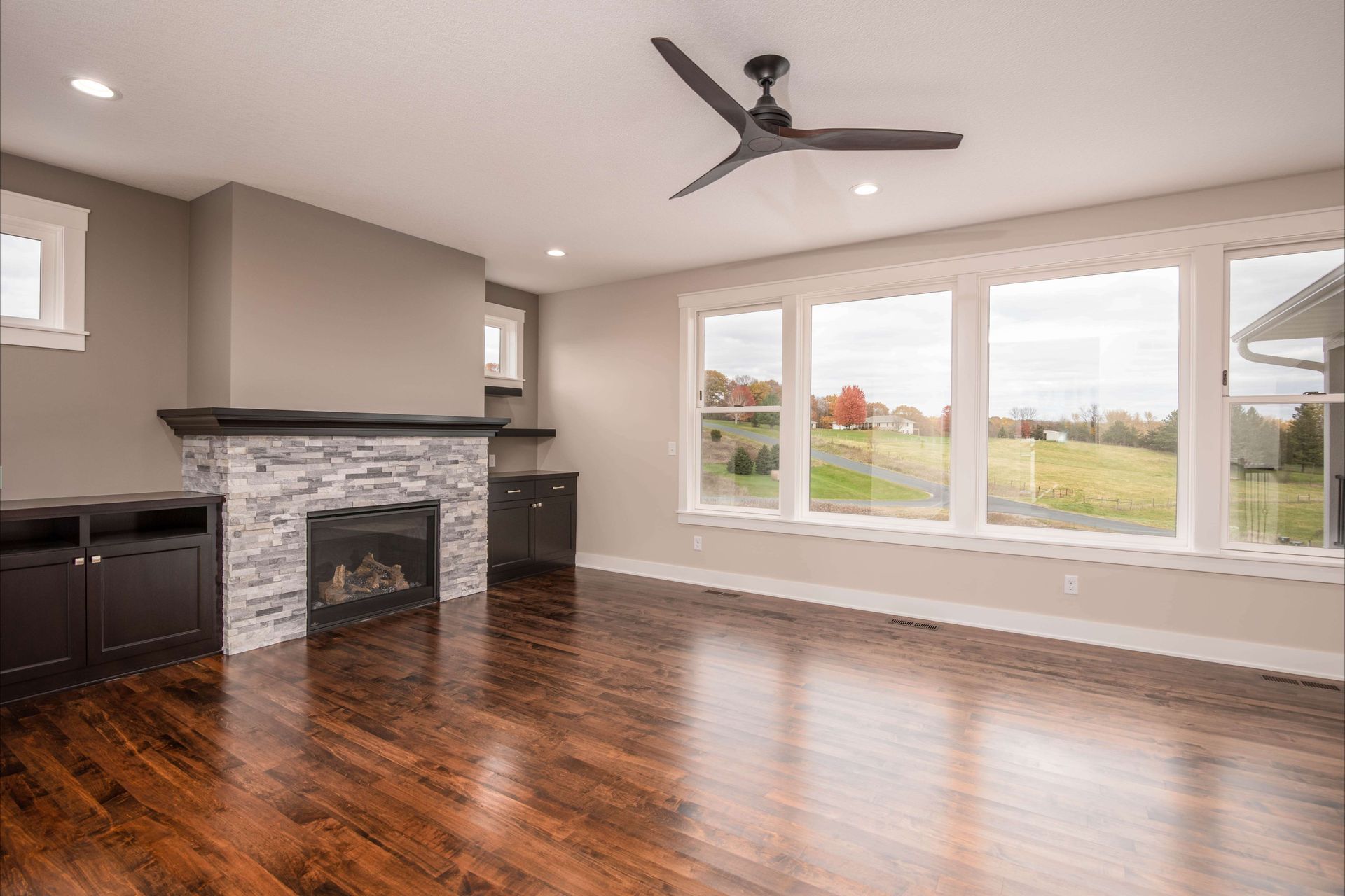 Living room with fireplace, windows, and dark wood floors. Gray walls, black entertainment unit, and a ceiling fan.