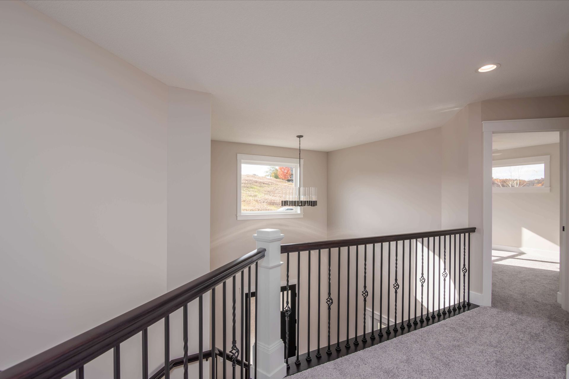 Hallway with brown railing, white walls, carpet flooring, window with view, and a hanging light fixture.