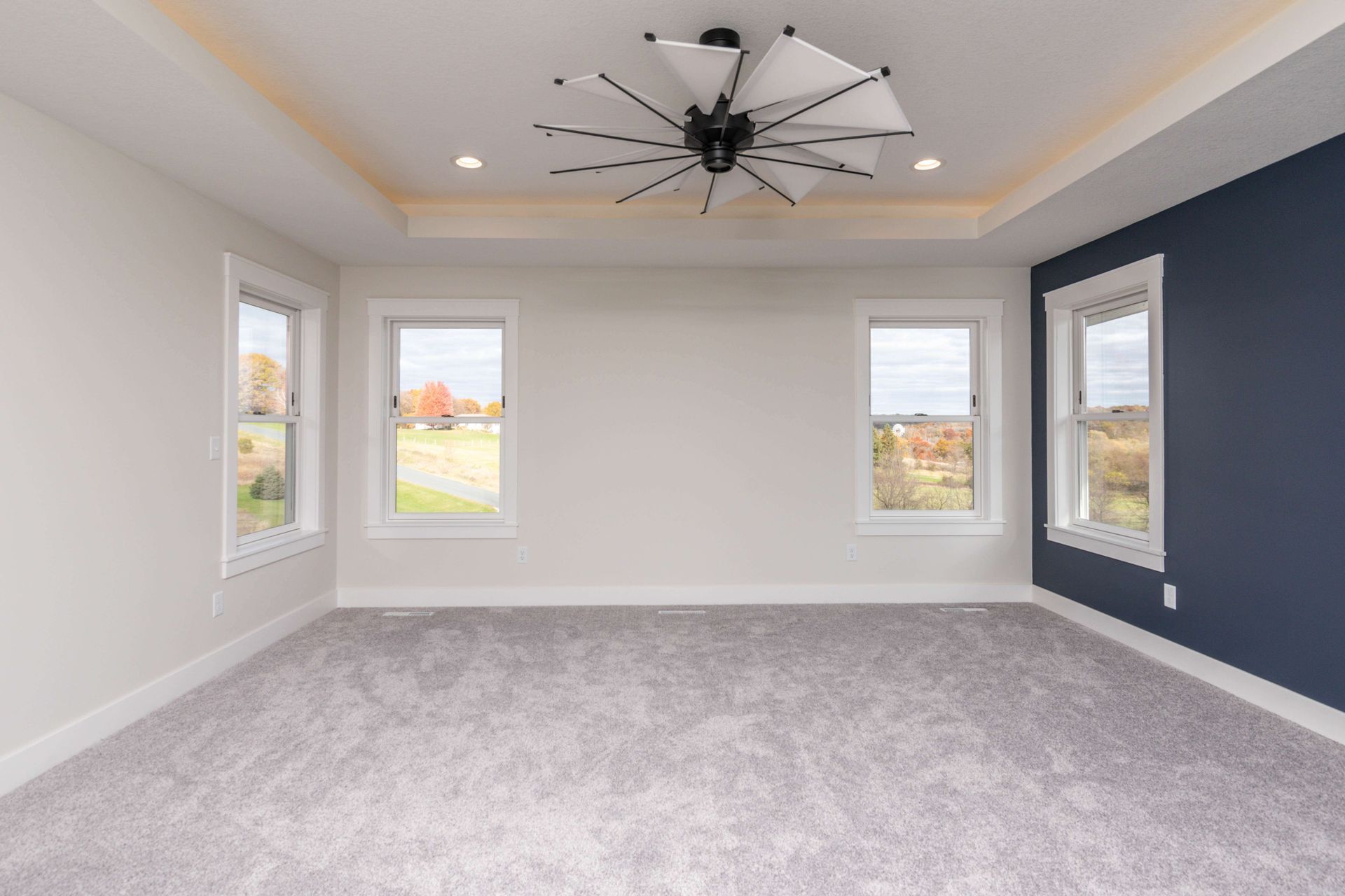 Spacious bedroom with gray carpet, white walls, and a navy accent wall. Features three windows, and a decorative ceiling fan.