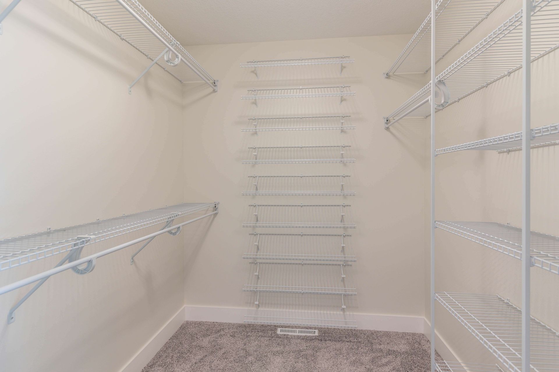 Empty walk-in closet with white wire shelving on three walls, beige walls, and carpet.