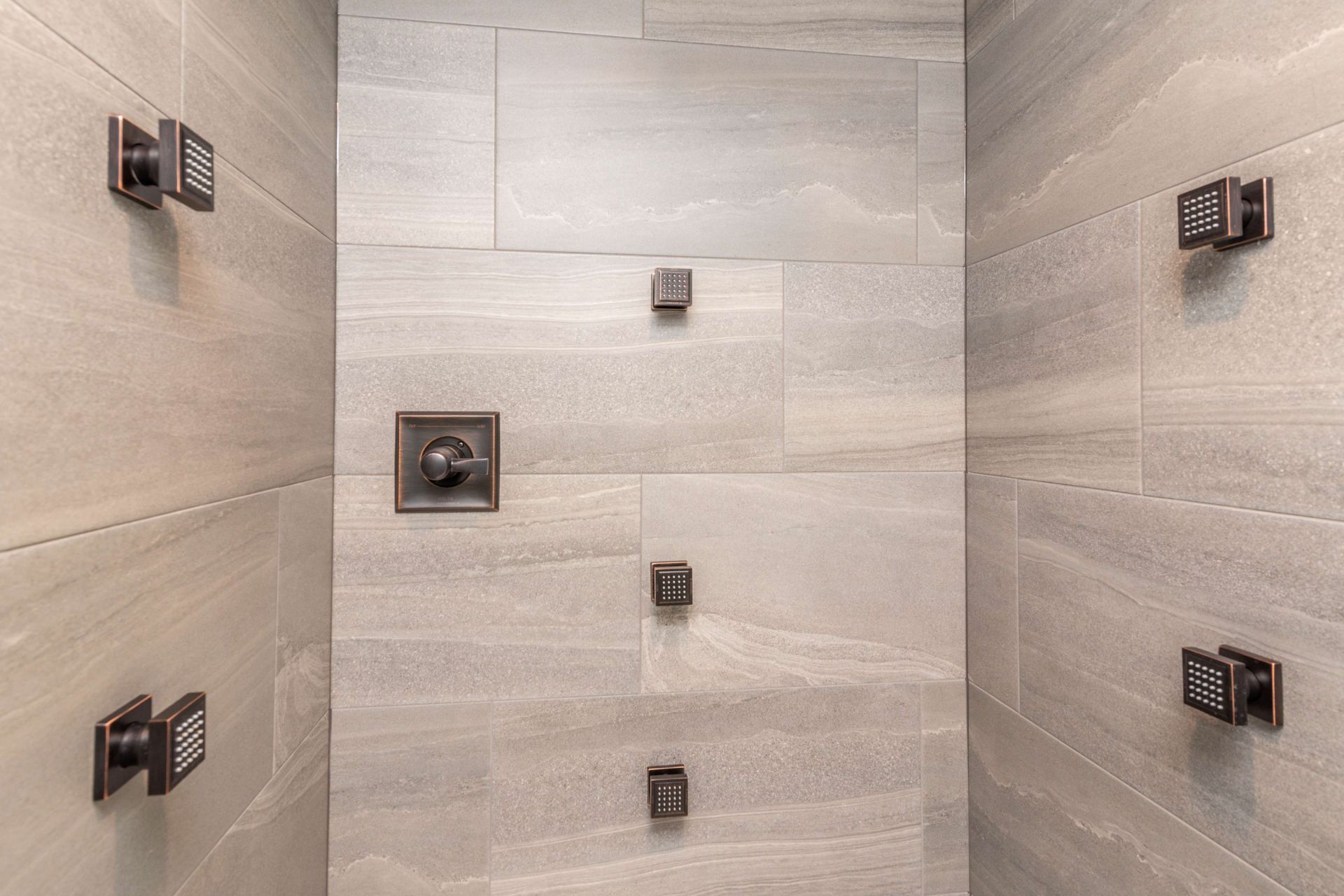 Gray tiled shower interior with multiple dark bronze water fixtures.