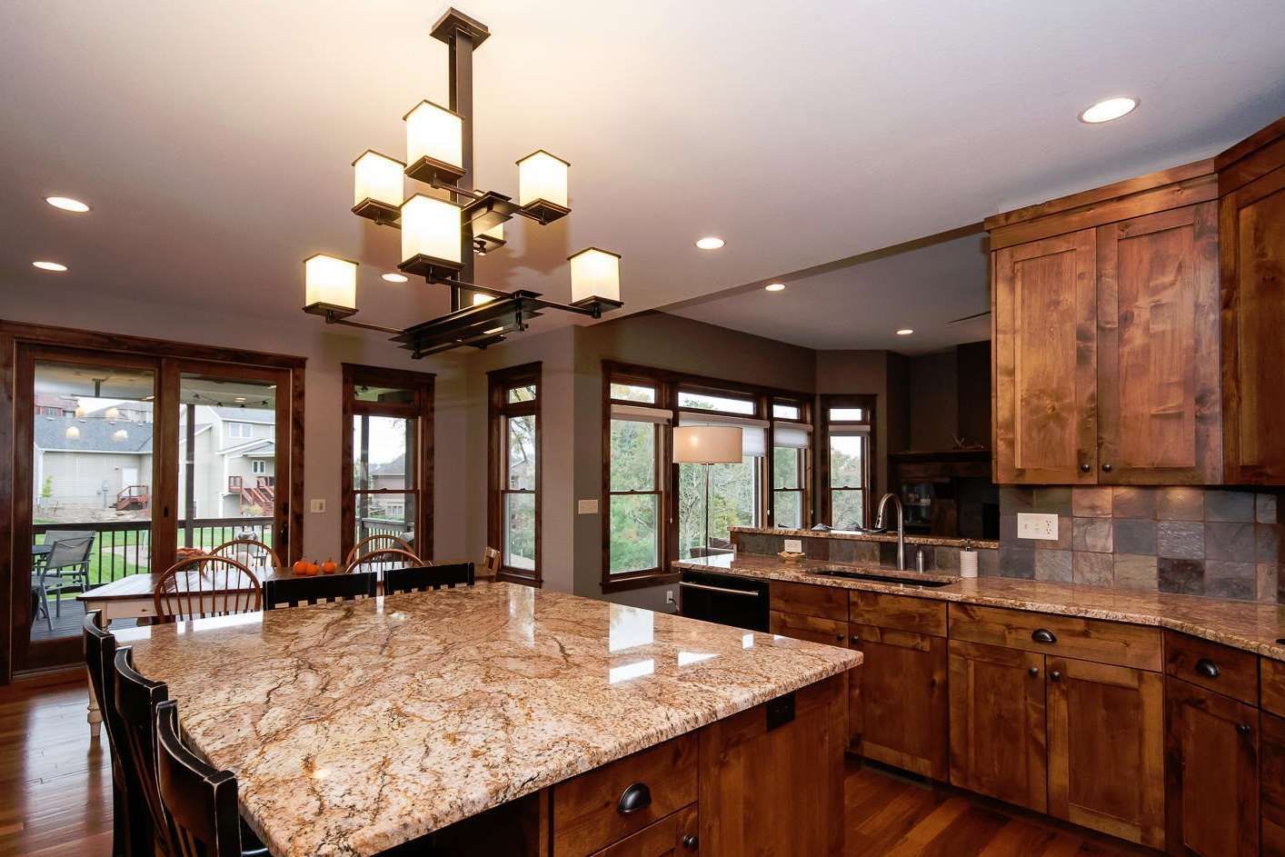 Kitchen with granite island, wooden cabinets, and large windows. Chandelier hangs above island.