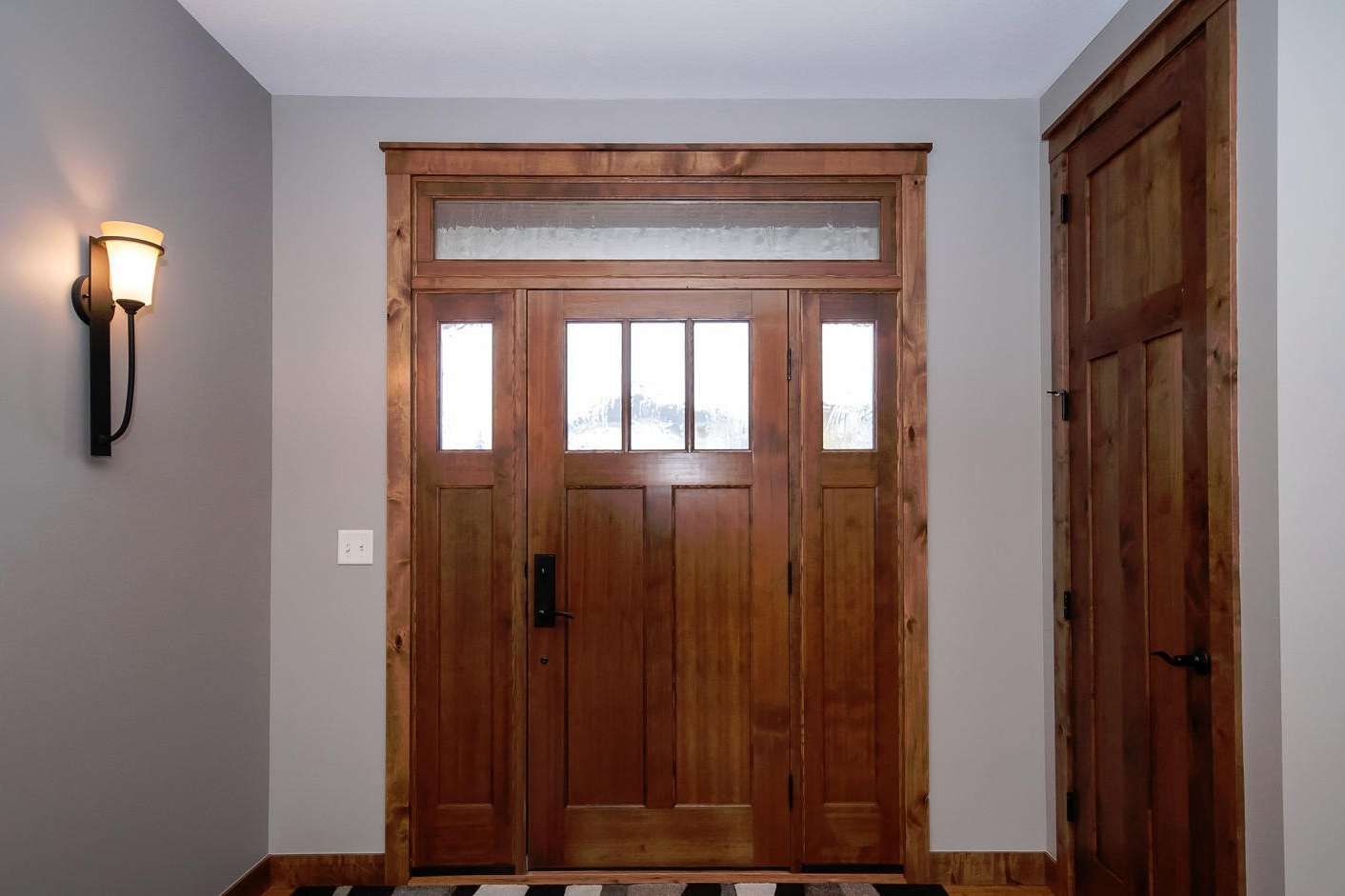 Wooden craftsman-style entryway with sidelight and transom window, next to a matching door and a wall-mounted sconce.