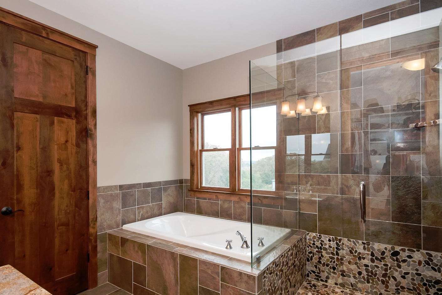 Bathroom with a white tub, stone tile walls, and a glass shower. Dark wood door and window frame.