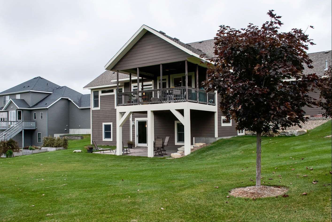 Two-story brown house with a deck overlooking a grassy yard; another house is to the left under an overcast sky.