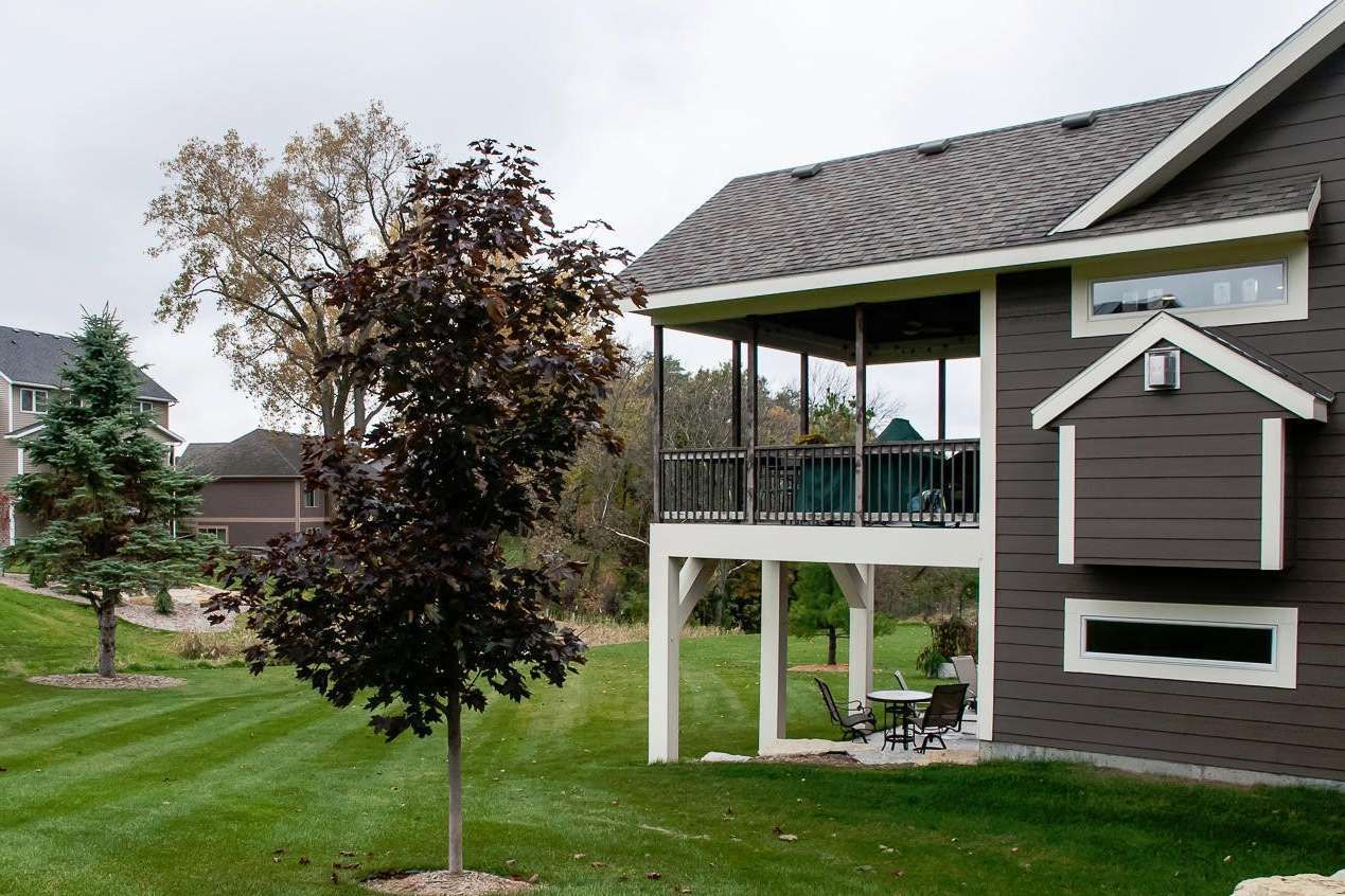 Backyard with a brown house, elevated screened porch, green lawn, trees, and cloudy sky.
