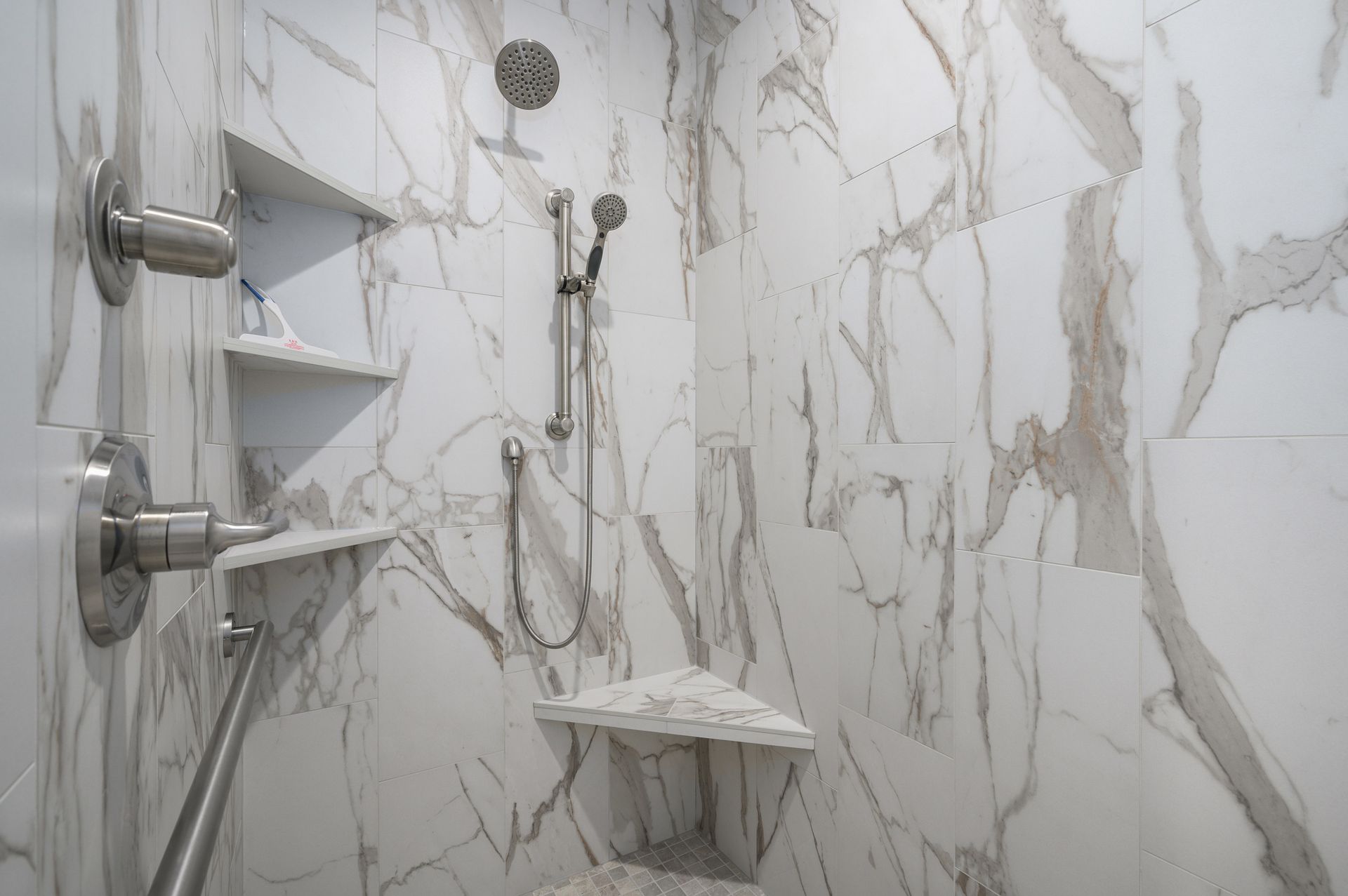 Shower with marble-look tile, built-in shelves, and accessible handrail. Chrome fixtures are installed.