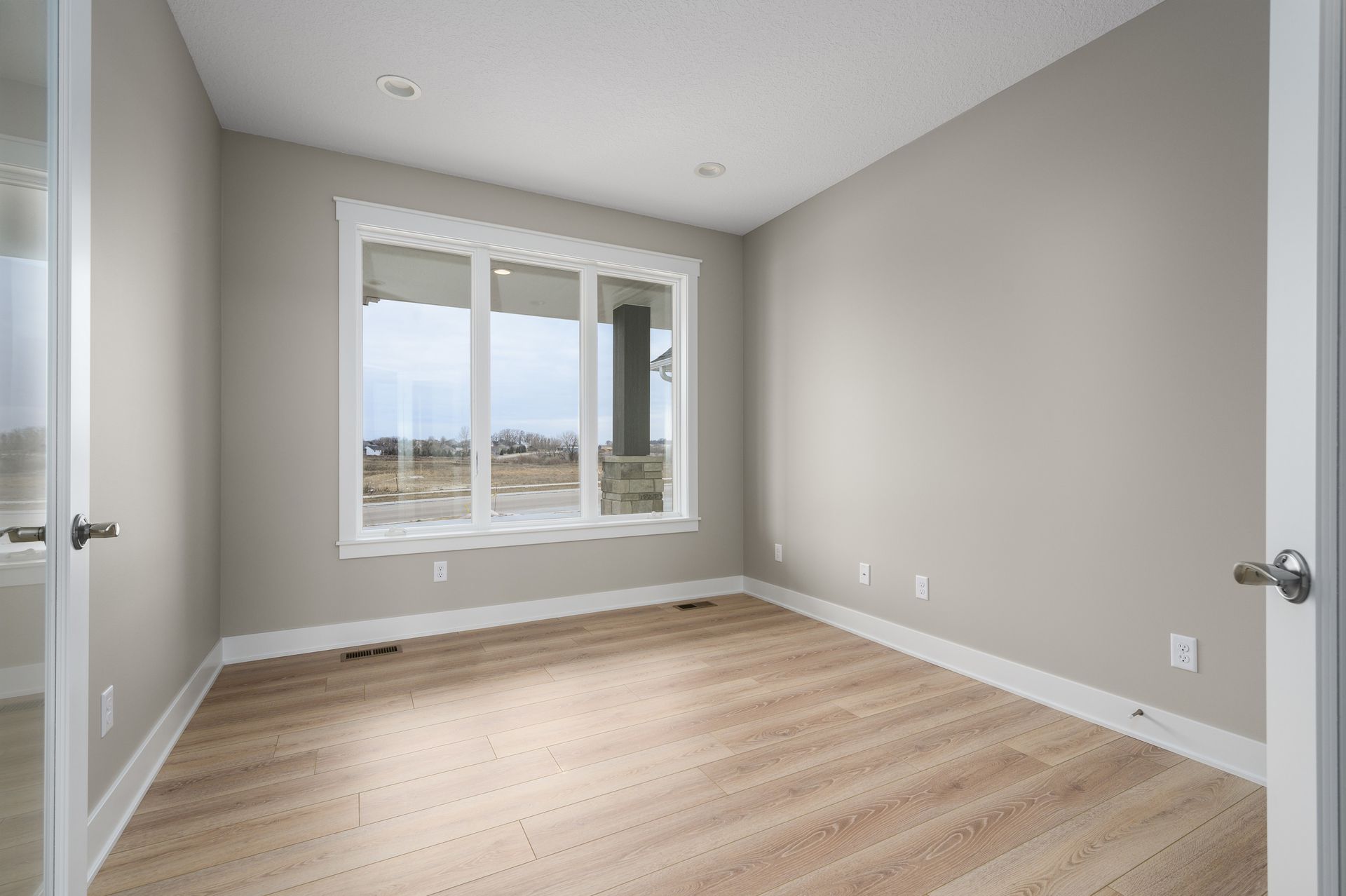 Empty room with large window, beige walls, light wood flooring, and open door.