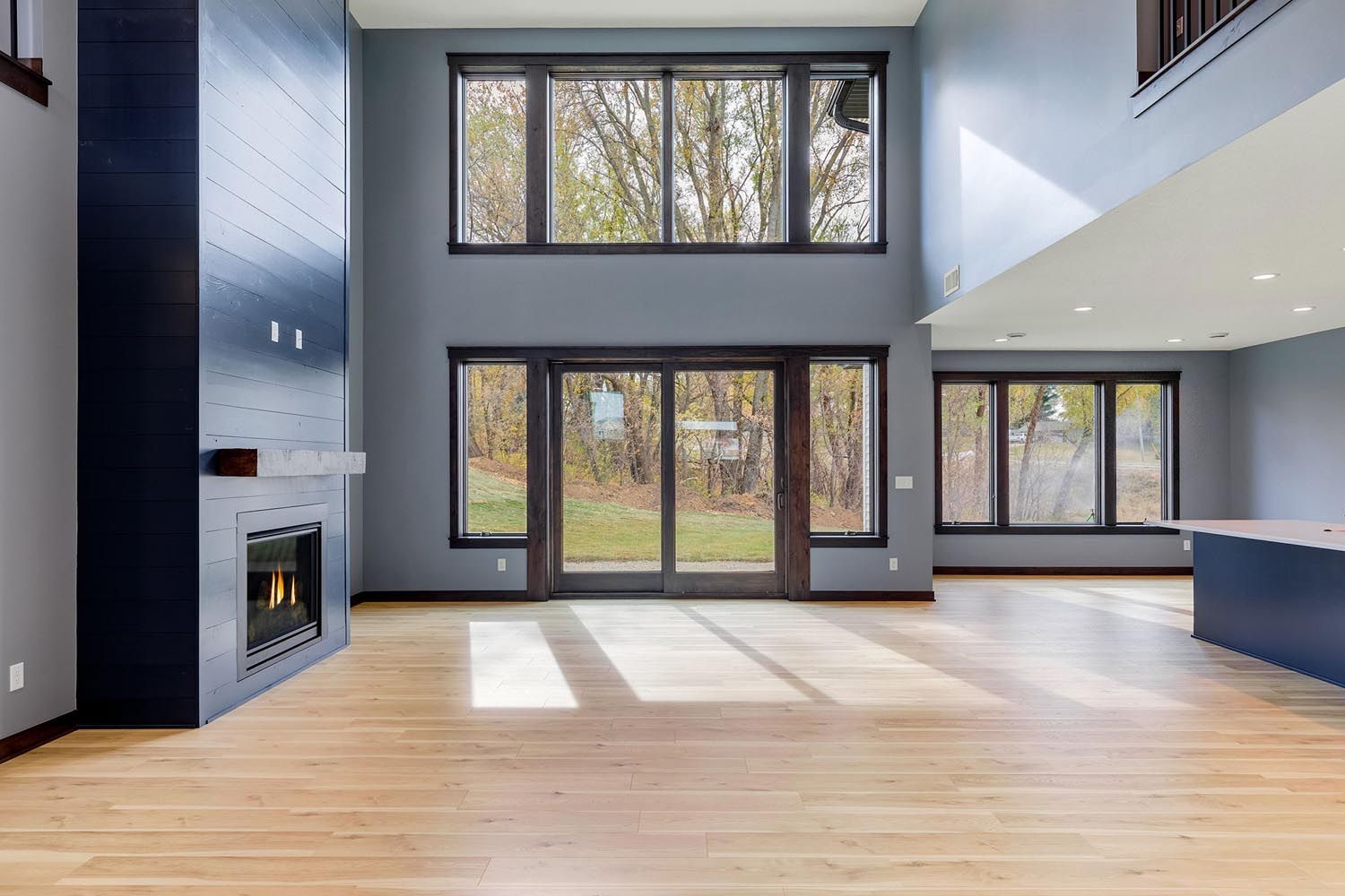 Spacious living room with large windows, fireplace, and hardwood floors. Gray walls and natural light.