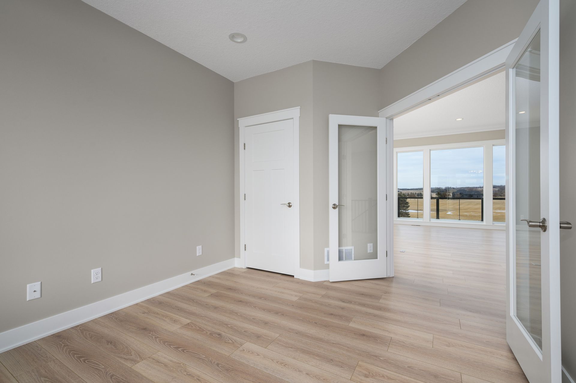 Empty room with light gray walls, wooden floor, and glass door leading to a room with large windows.