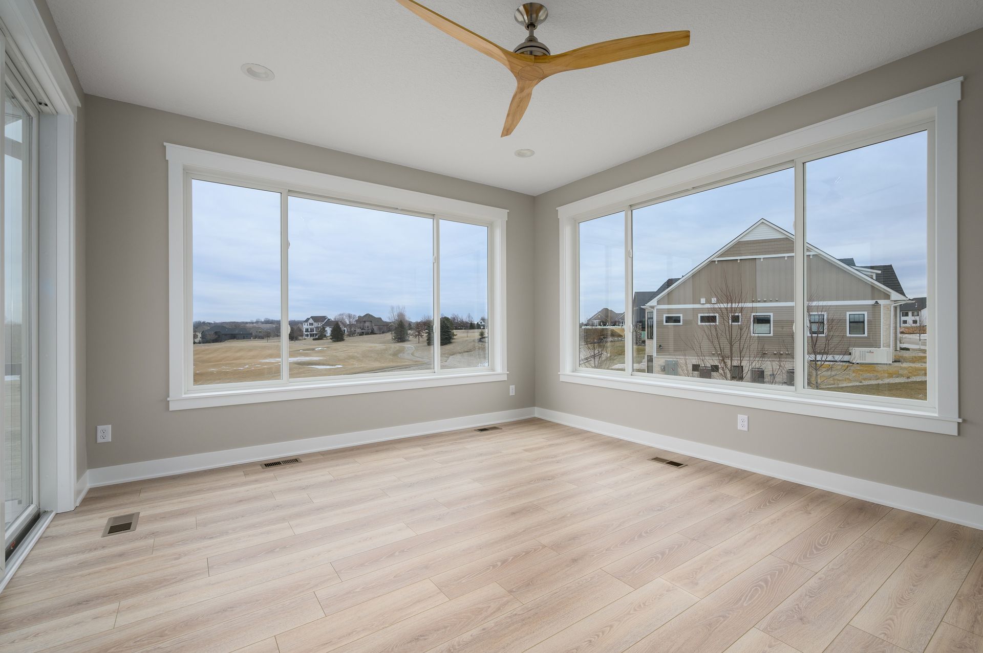 Empty room with large windows, wood floors, and a ceiling fan, overlooking a suburban landscape.