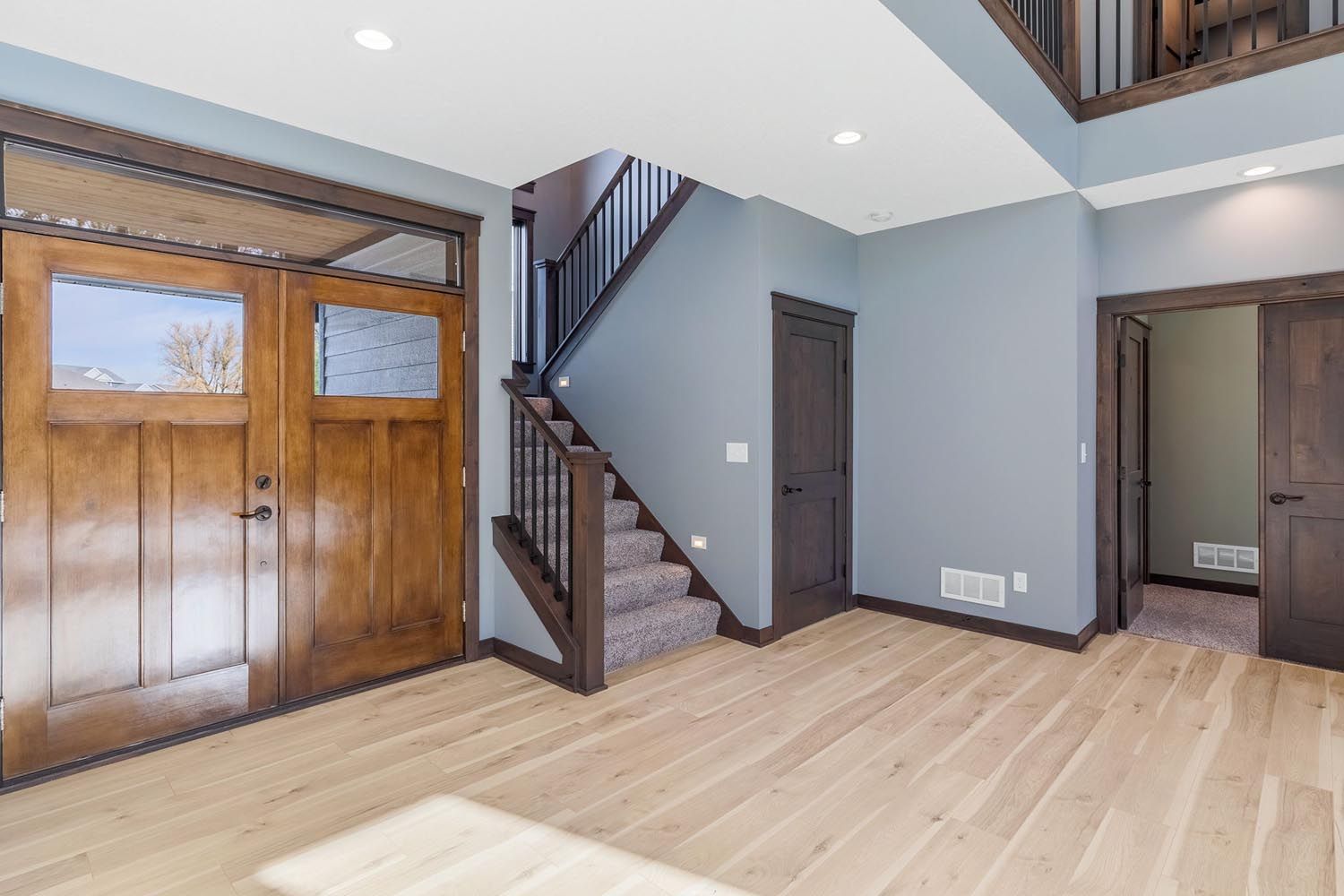 Entryway with wooden doors, stairs, and light wood floors, blue-gray walls, and dark brown trim.