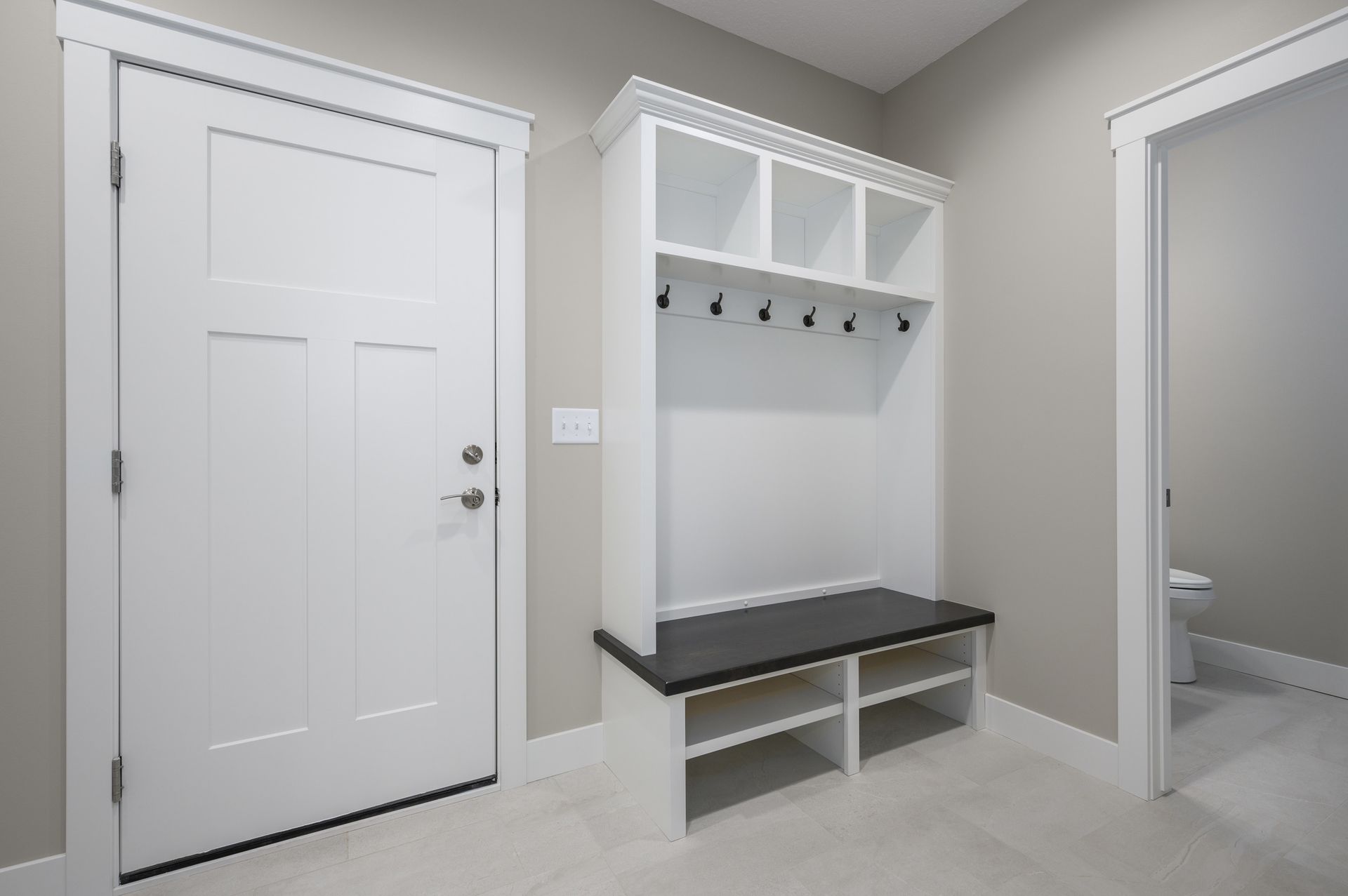 Mudroom with white door, built-in storage bench, and open shelving, neutral walls, and a glimpse of a toilet.