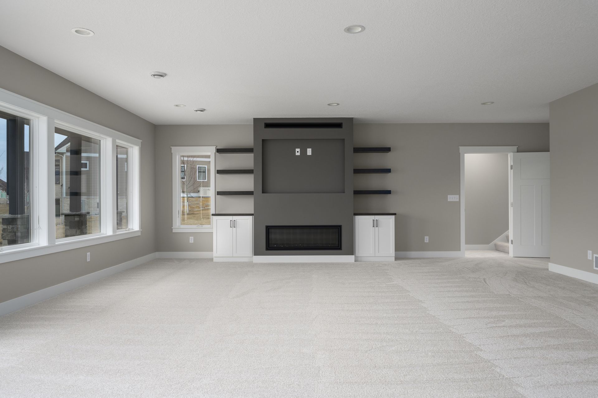 Empty living room with fireplace, built-ins, large windows, and neutral carpet and walls.