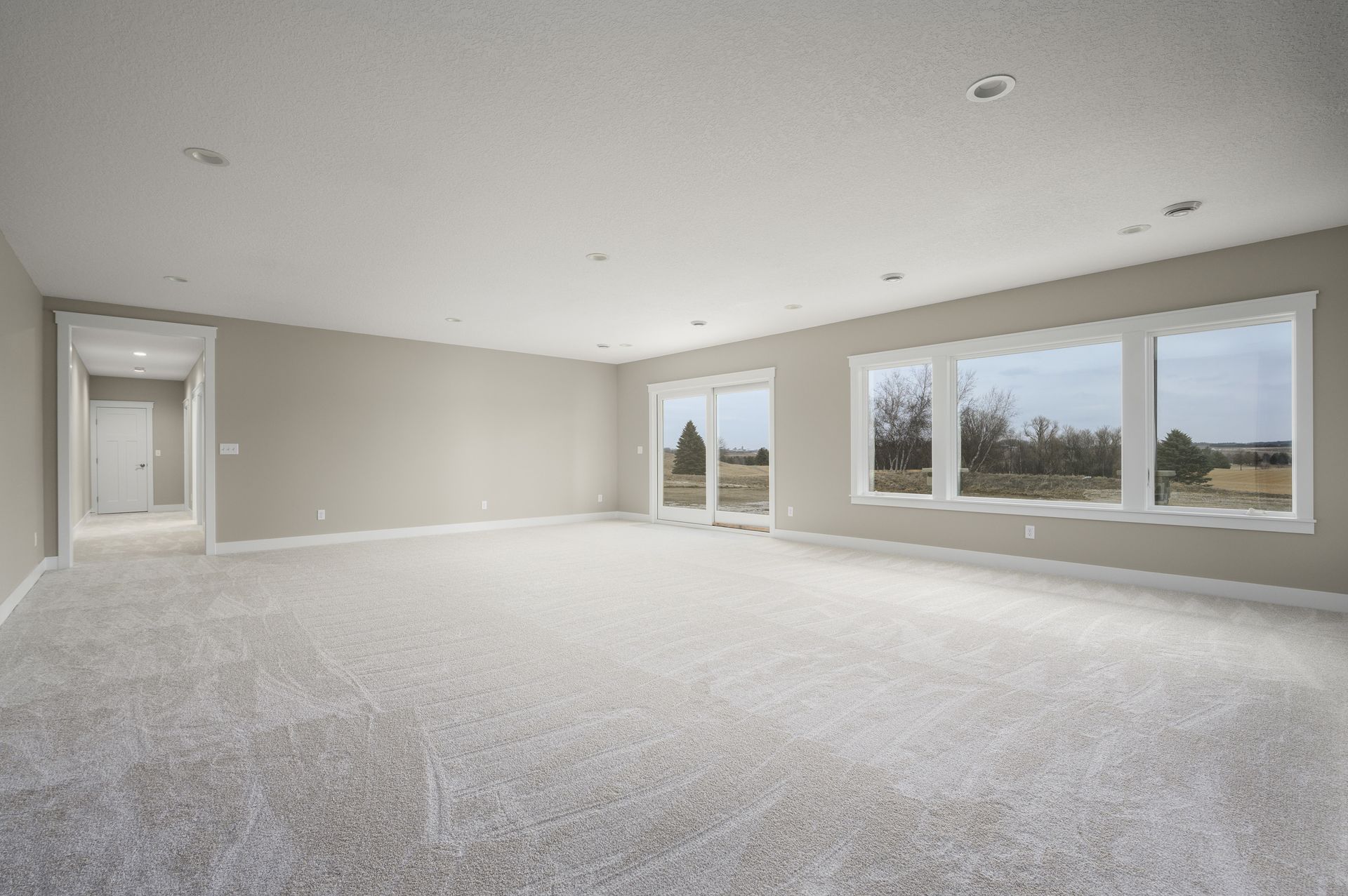 Empty room with light-colored carpet, walls, and ceiling. Large windows and sliding glass door provide natural light.