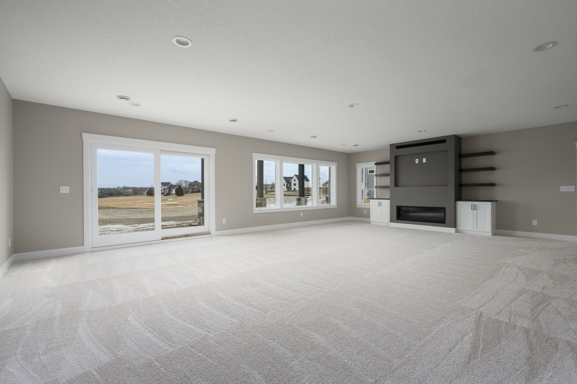 Empty living room with light gray walls and carpet, large windows, and a fireplace.