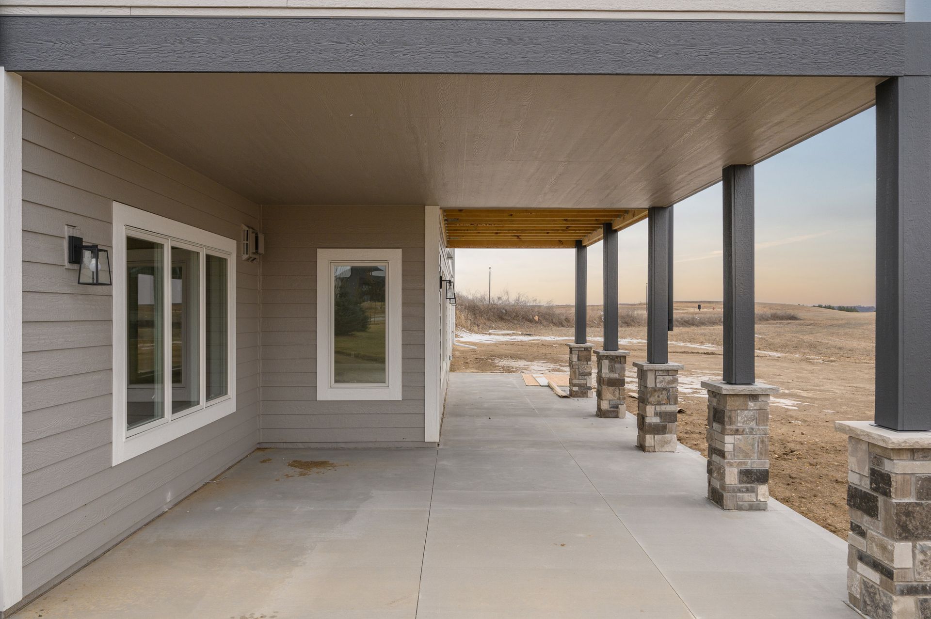 Covered porch with gray siding, windows, and dark columns; open landscape in the background.
