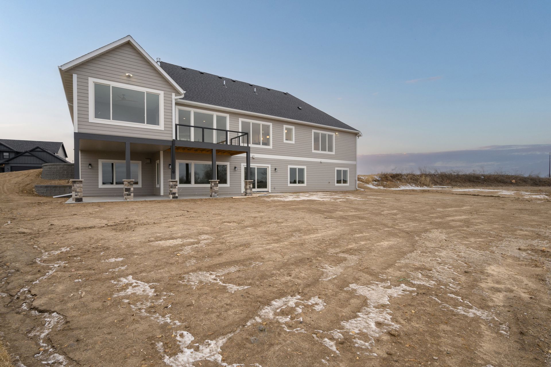 Two-story house with gray siding and a deck, in a vacant dirt lot under a blue sky.