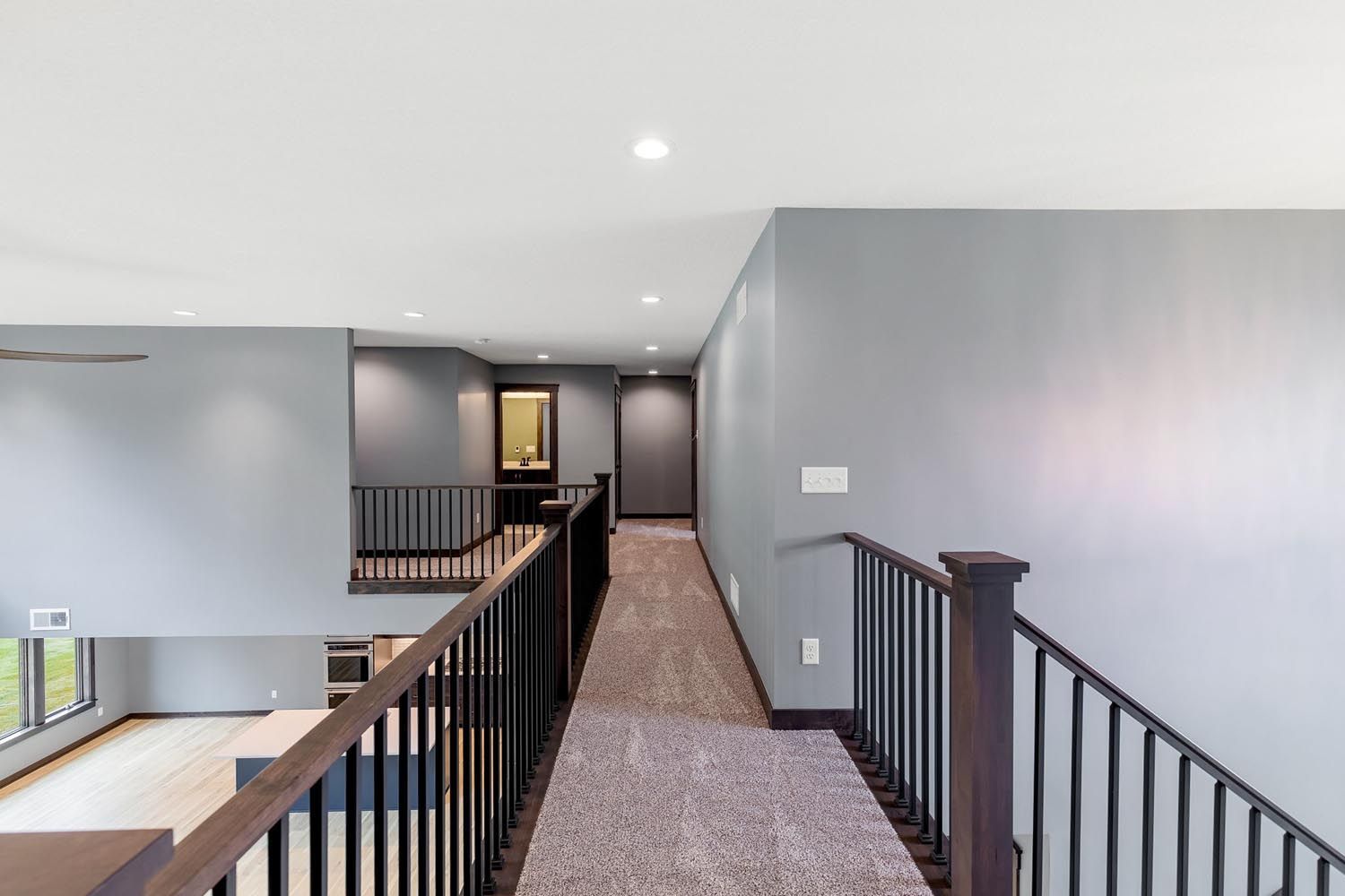 Hallway with gray walls, carpet, and black railing. Doorways and recessed lighting.