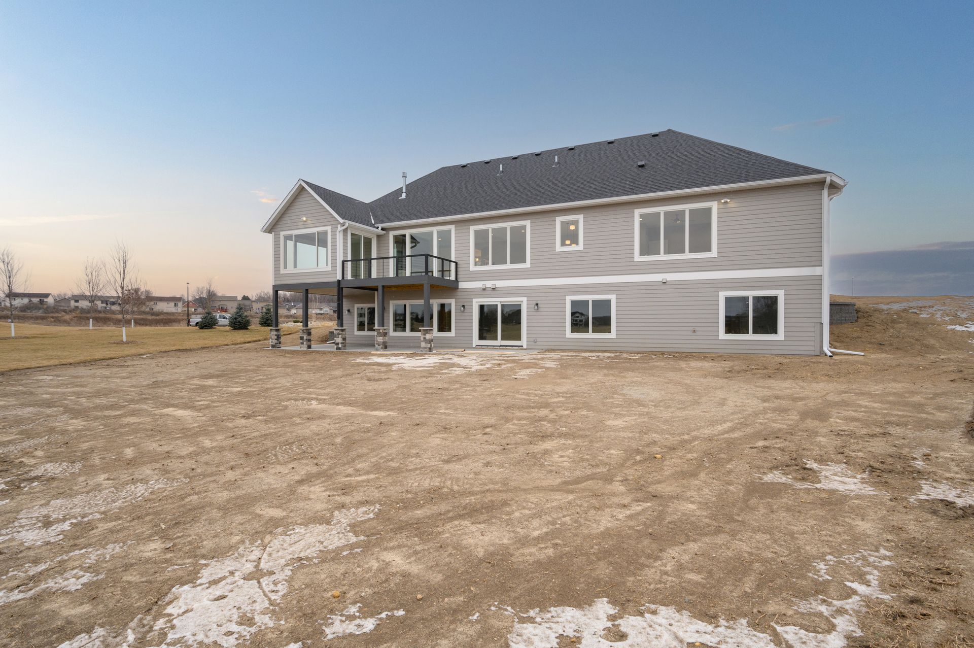Two-story house with light gray siding, deck, and large windows on a barren lot under a blue sky.