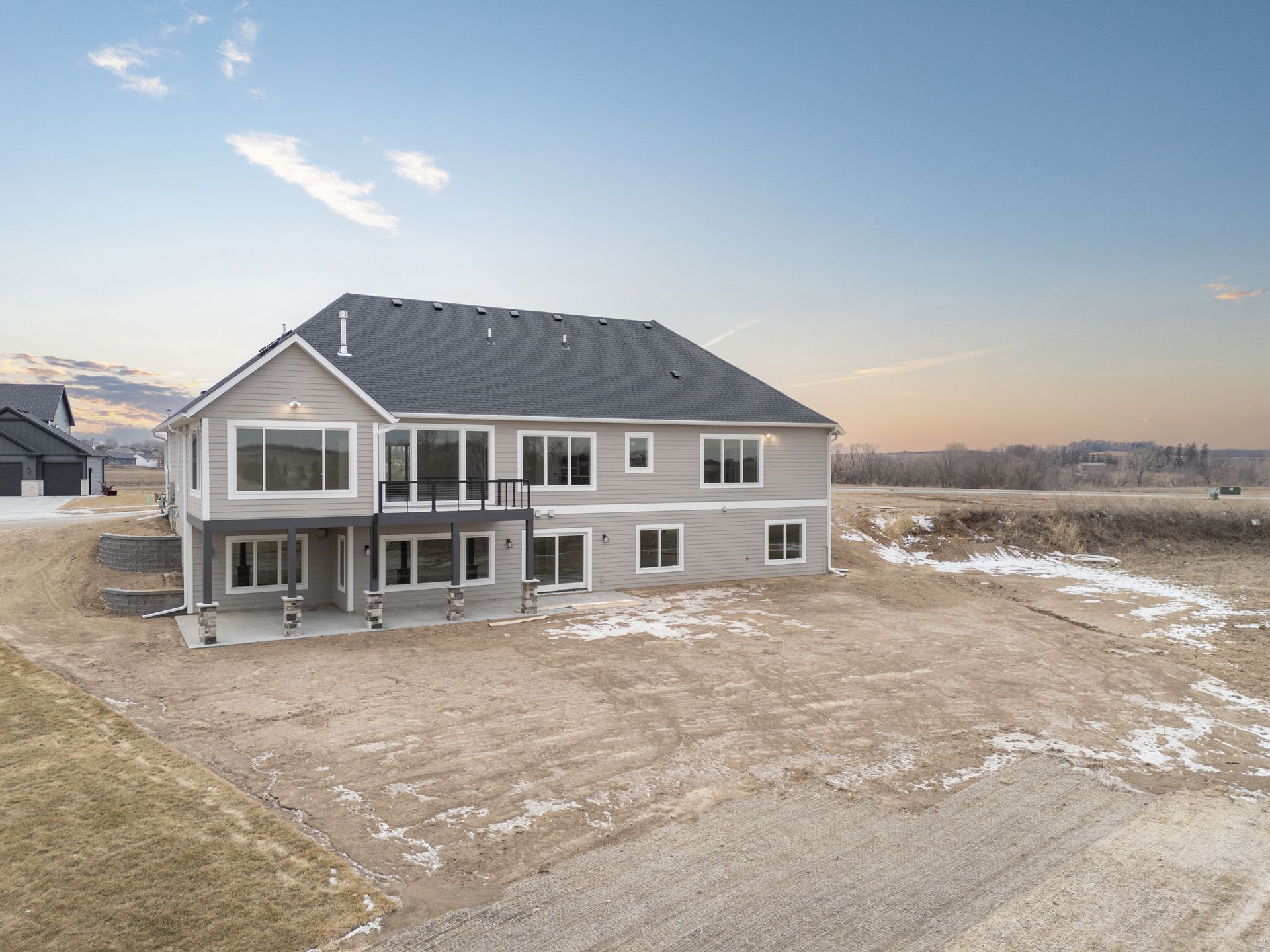 Gray two-story house with a deck in a sparsely populated area under a partly cloudy sky.