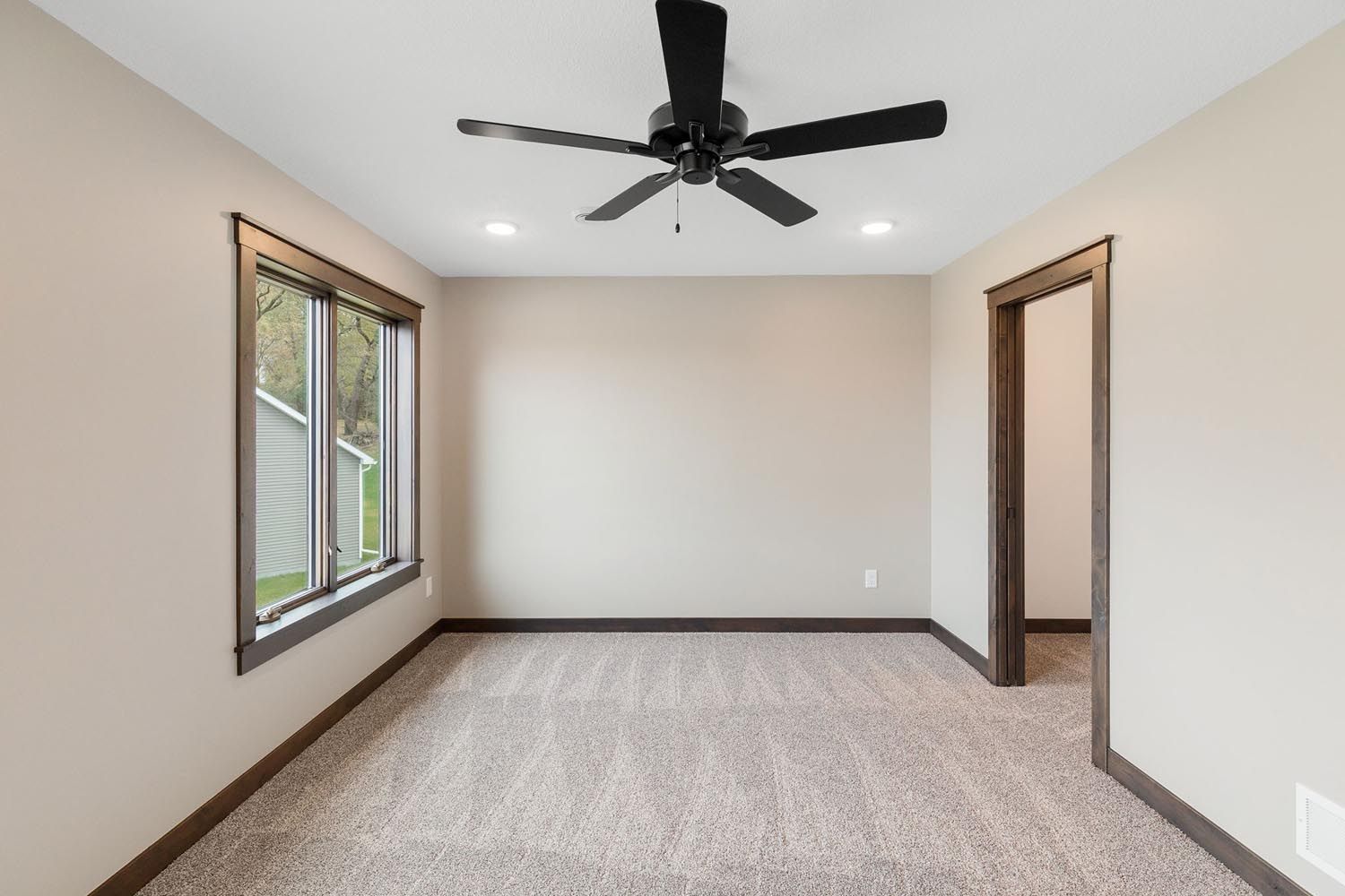 Empty room with tan walls, brown trim, carpet, ceiling fan, window, and doorway.