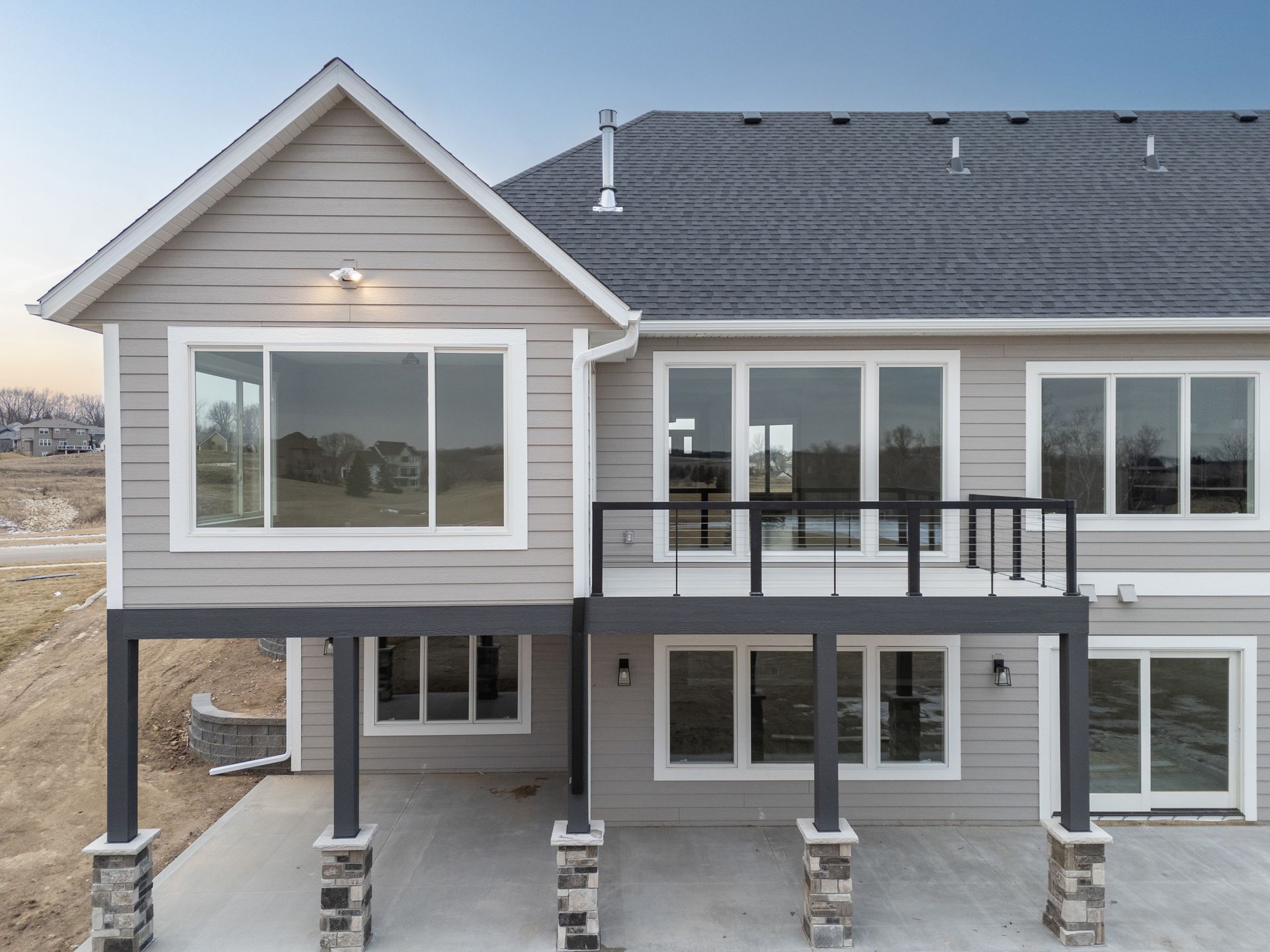 Back of a two-story beige house with a deck. Large windows, gray roof, dark posts with stone bases.