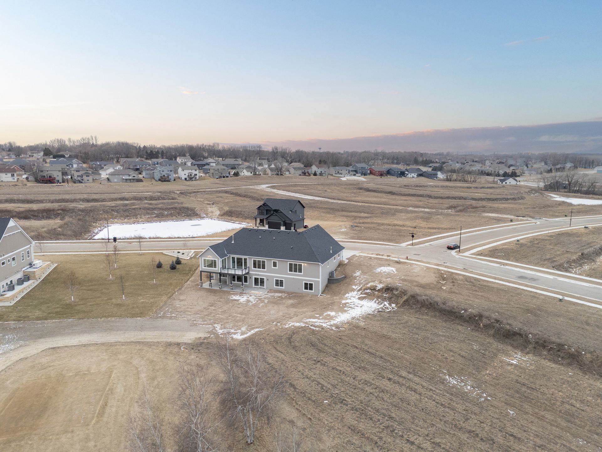 Aerial view of a gray house in a new development, with a road and barren landscape under a pale sky.