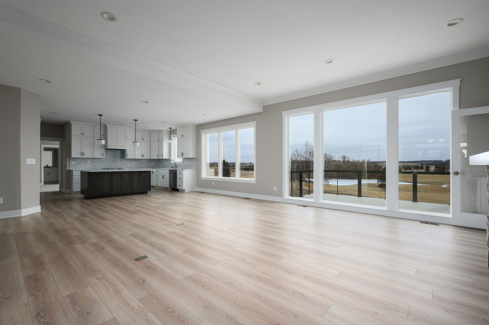 Open-concept living space with large windows, light wood floors, and a kitchen with white cabinets.