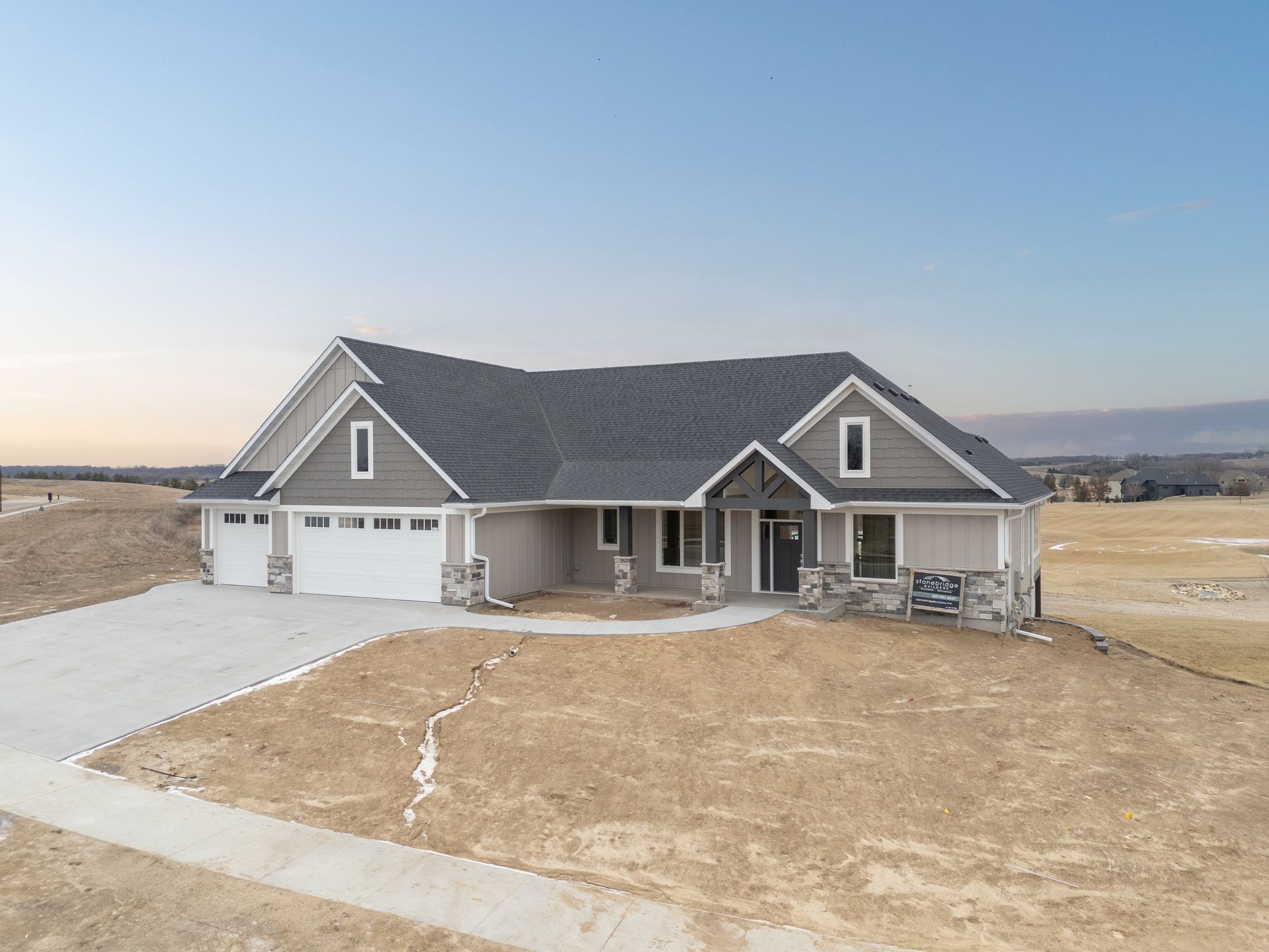 Exterior view of a new gray house with a three-car garage and light brown siding on a vacant lot.