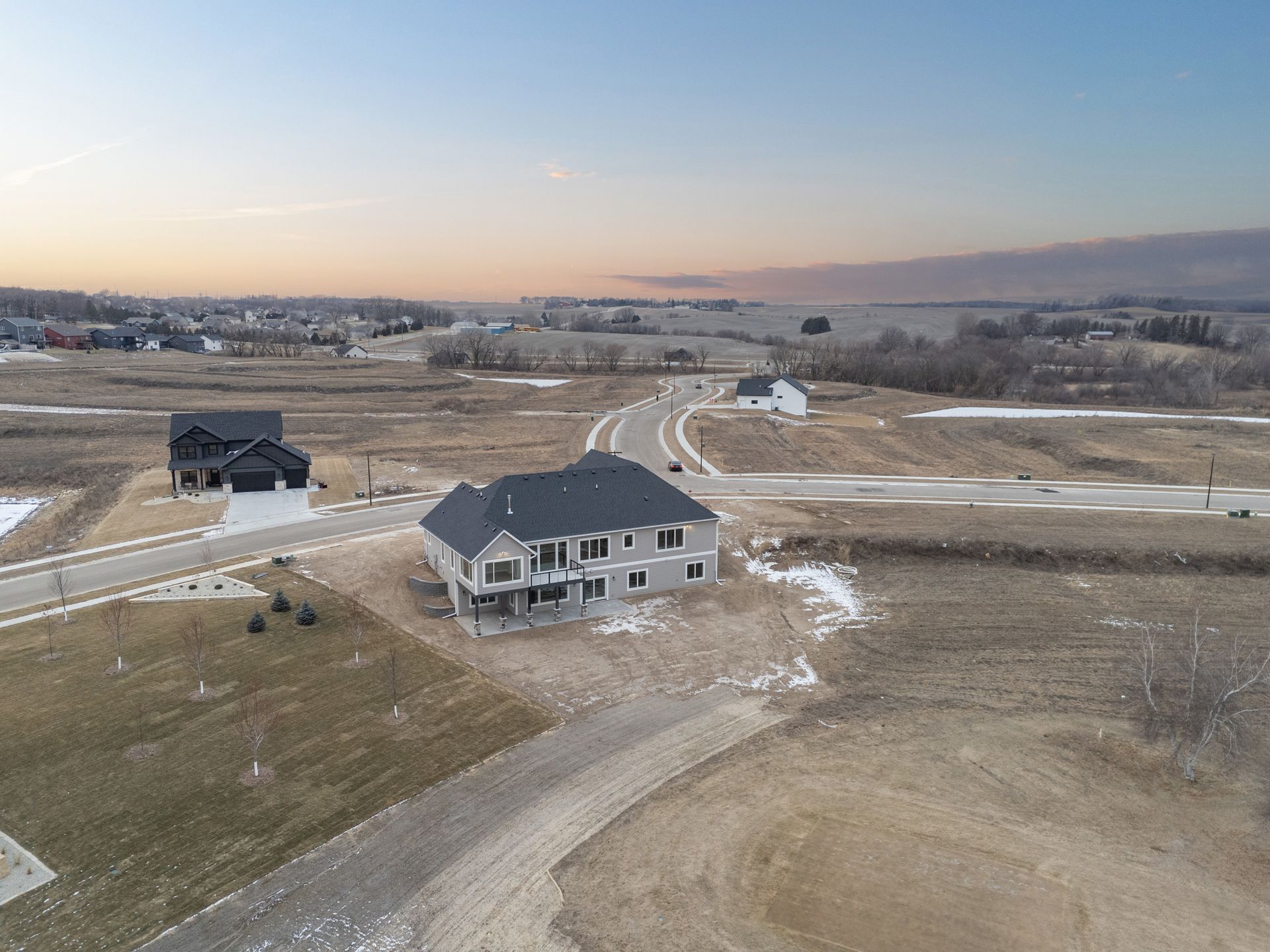 Aerial view of two-story gray house with dark roof in a new residential development. Brown fields and light blue sky.