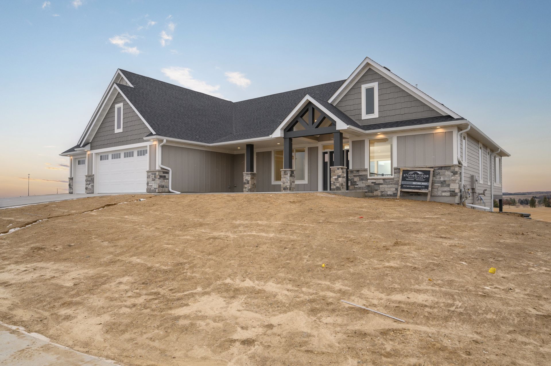 A light gray house with a black roof, stone accents, and a barren yard under a blue sky.