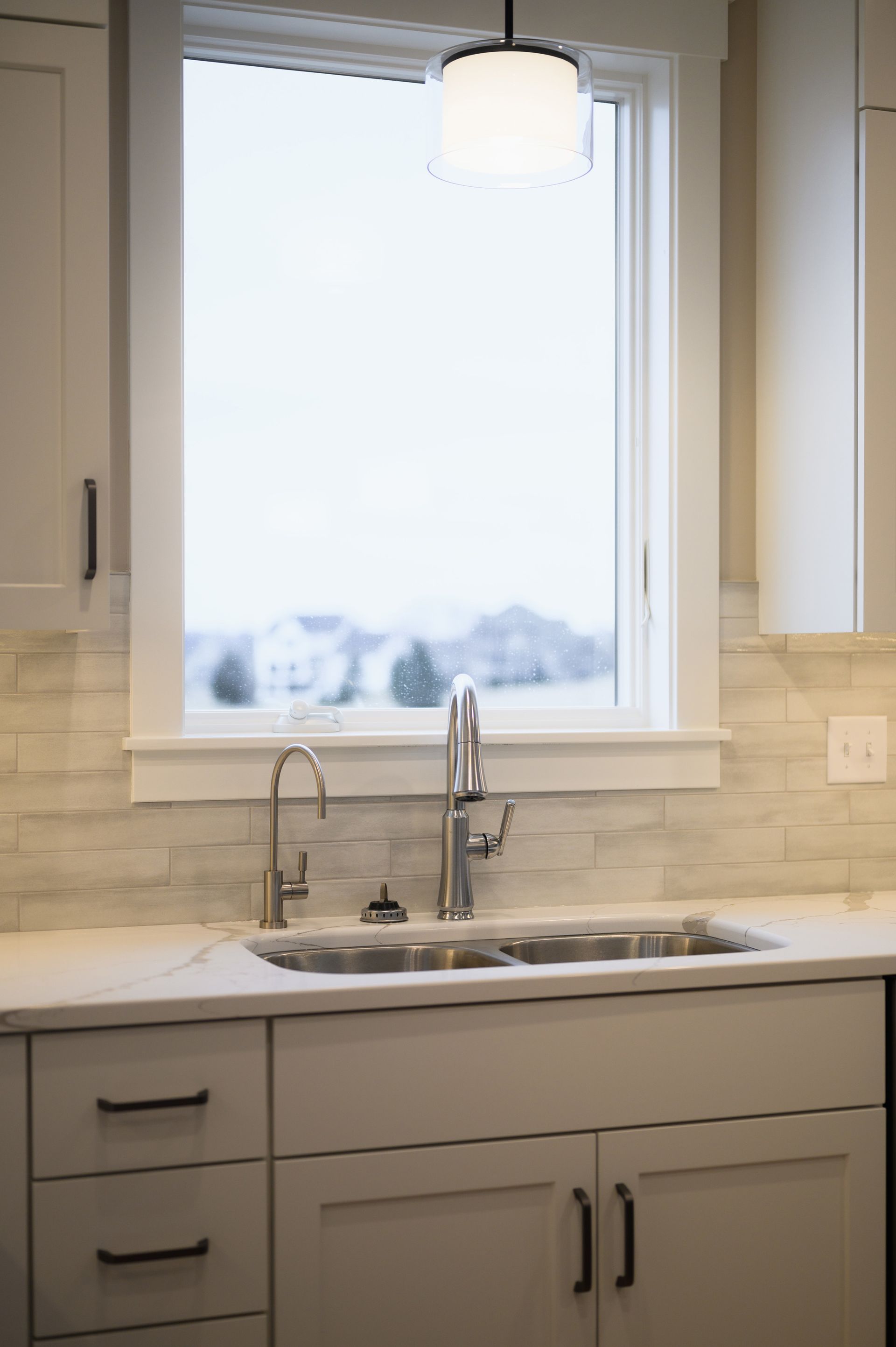 Kitchen sink with window above, white cabinets, marble countertop, faucet and light fixture.