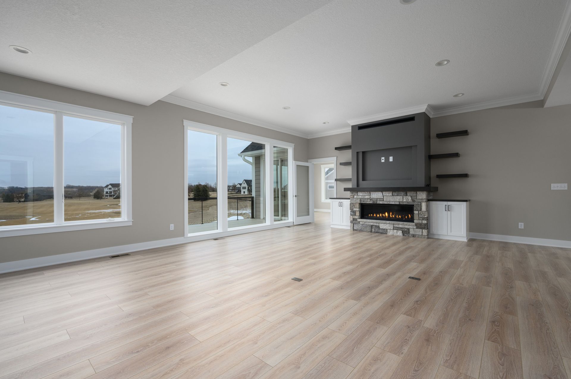 Spacious, empty living room with light wood floors, large windows, fireplace, and built-in shelving.