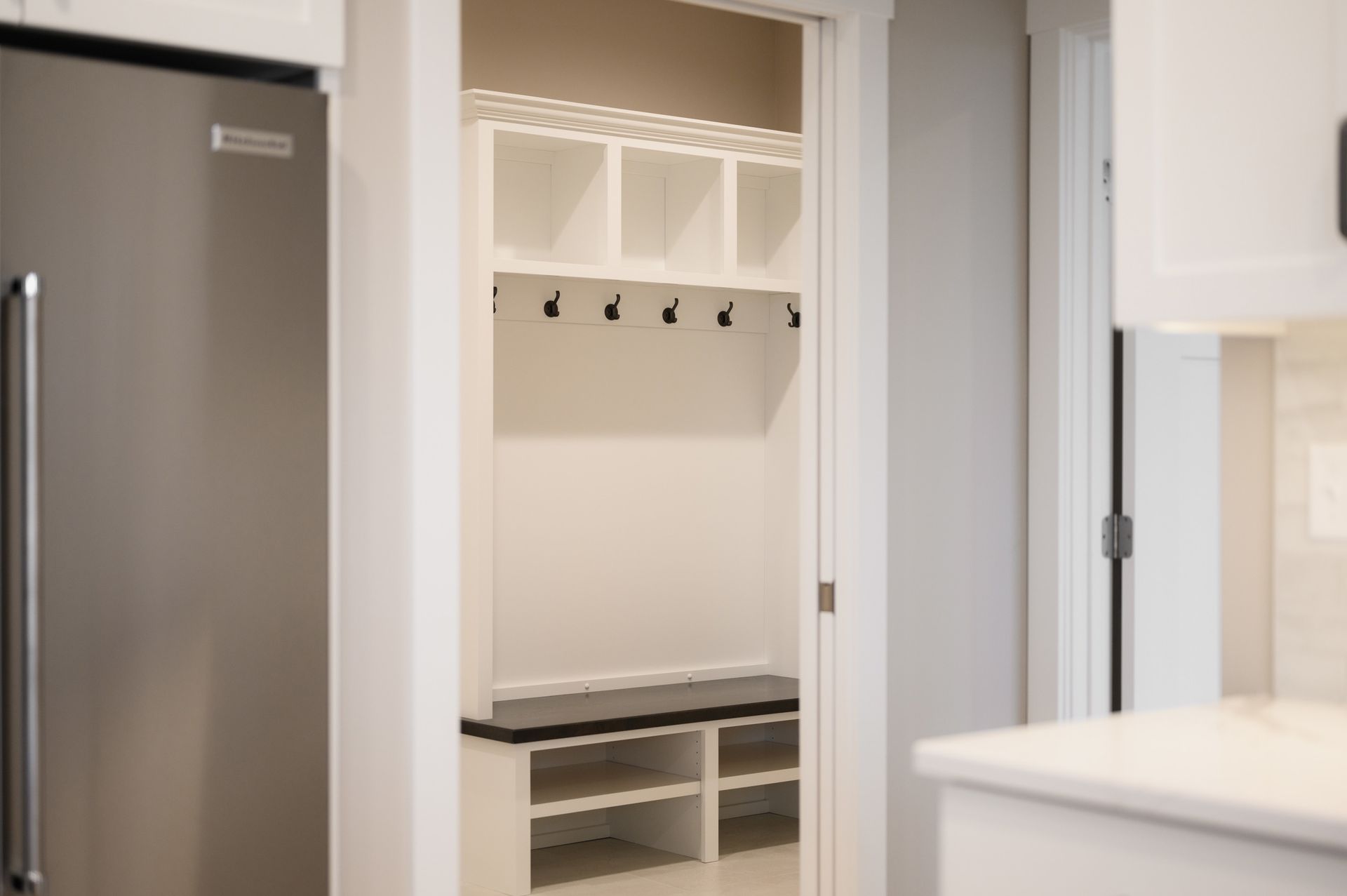 White entryway with shelving, hooks, bench, and shoe storage, next to a stainless steel refrigerator.