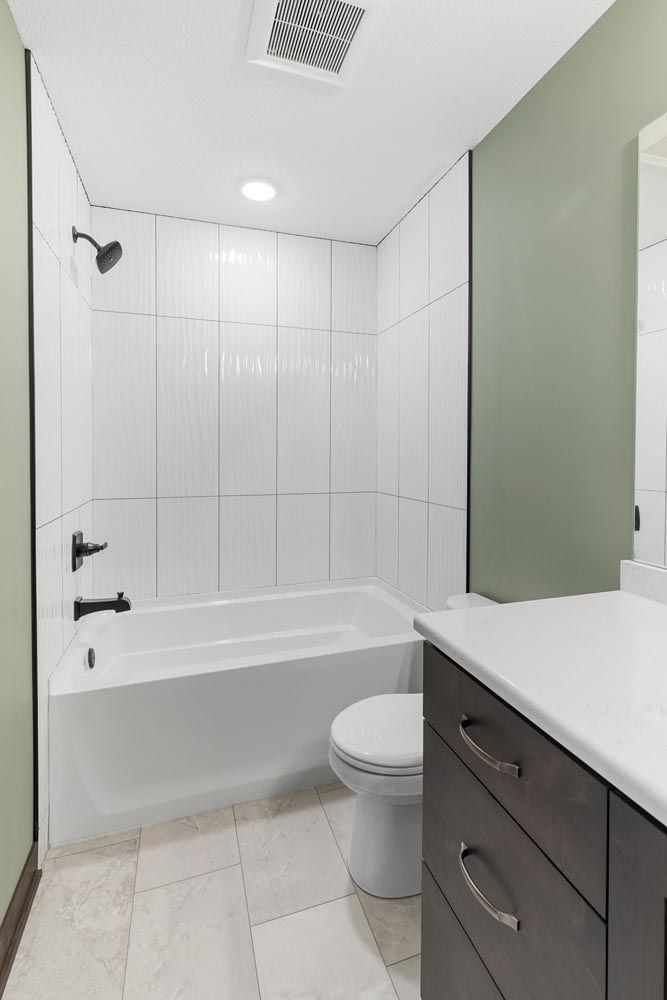 Bathroom with white tiled shower, tub, toilet, and vanity with dark cabinets and a white countertop.