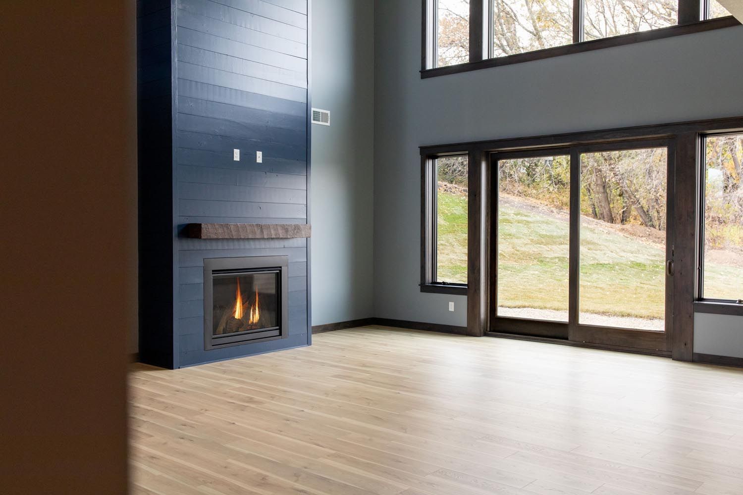 Empty living room with blue accent fireplace, light wood floor, and large windows overlooking a yard.