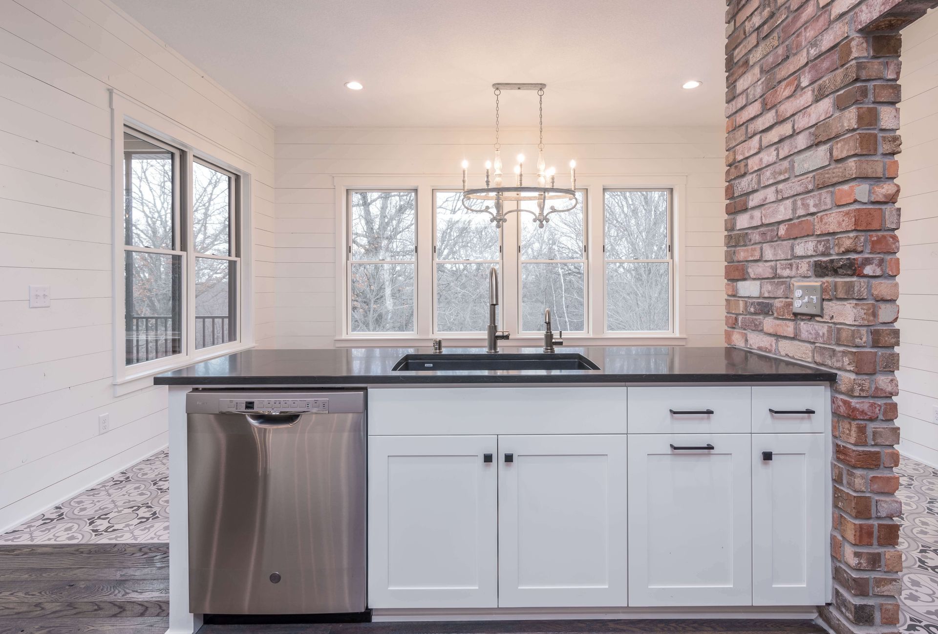 Modern kitchen island with stainless steel appliances, white cabinets, dark countertop, and exposed brick column.