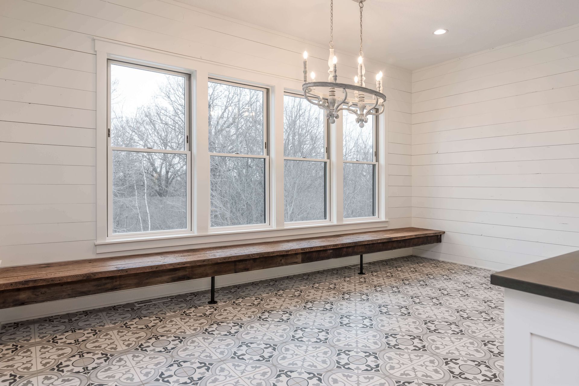 Bright dining room with large windows, wooden bench, chandelier, patterned tile floor, and white walls.
