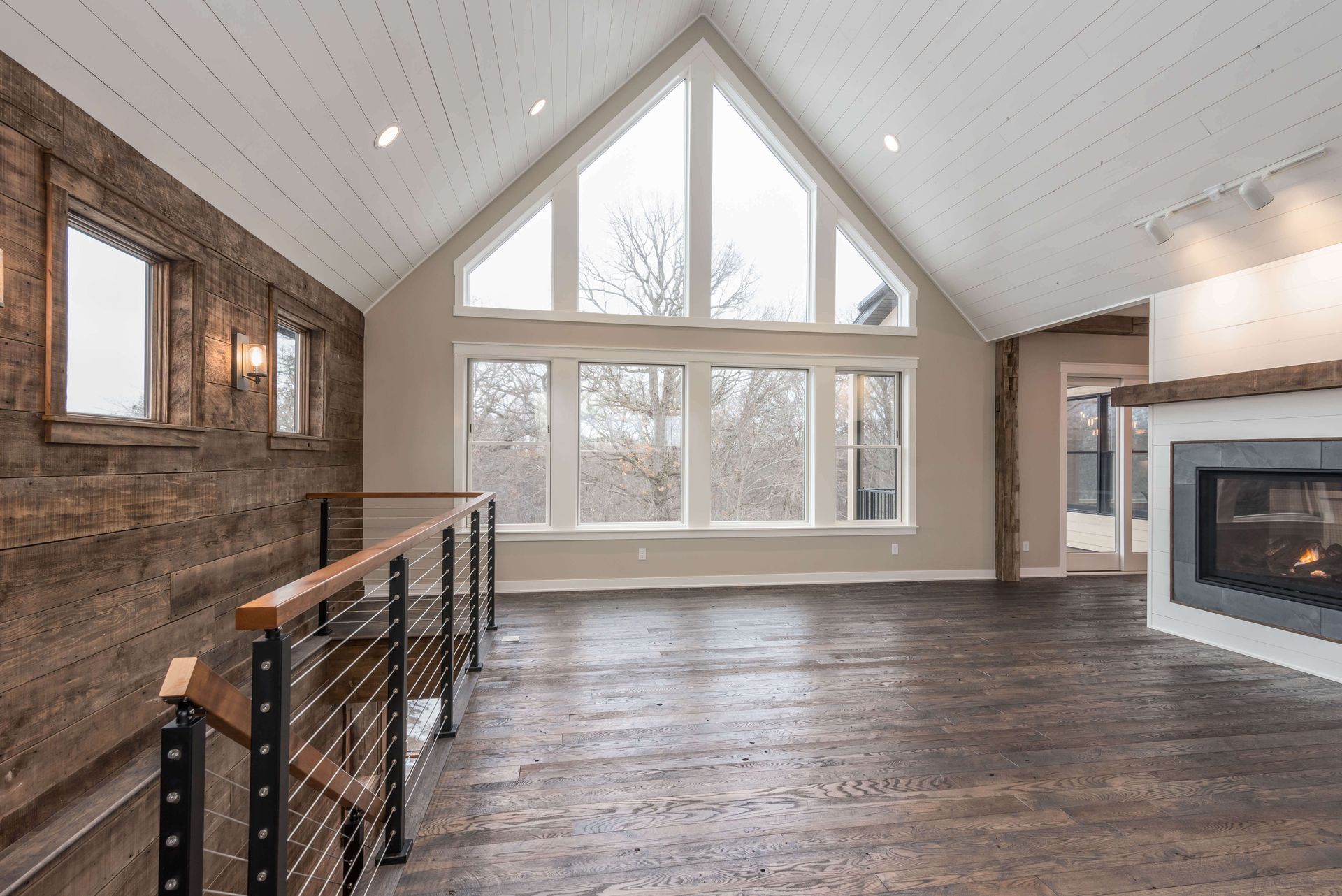 Spacious, light-filled room with large windows, fireplace, and exposed wood siding, overlooking an outdoor view.