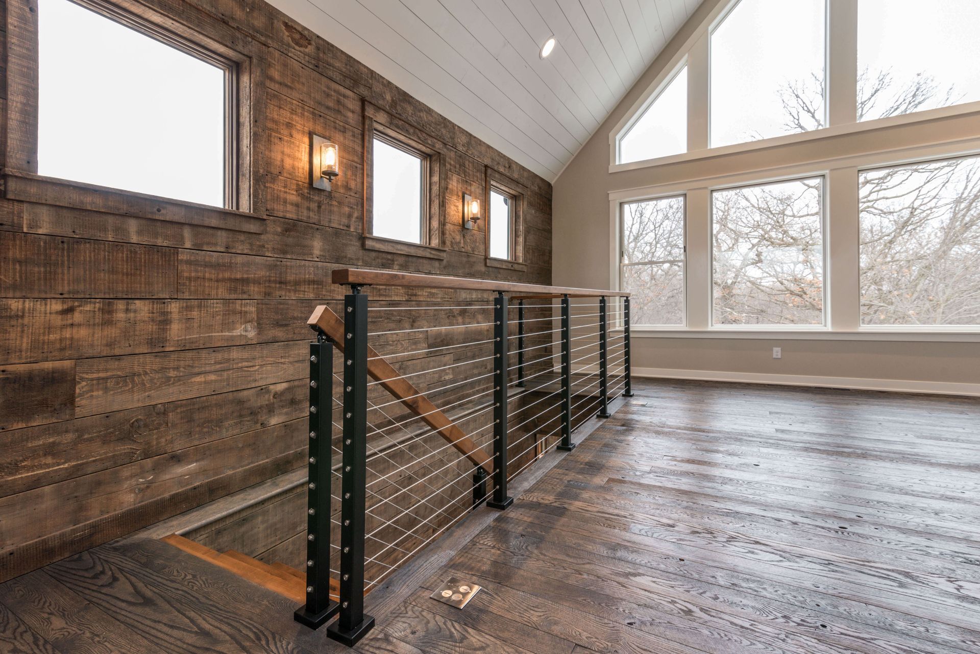 Interior view: Dark wood paneled wall, stairs with metal and cable railing, bright windows, and wood flooring.