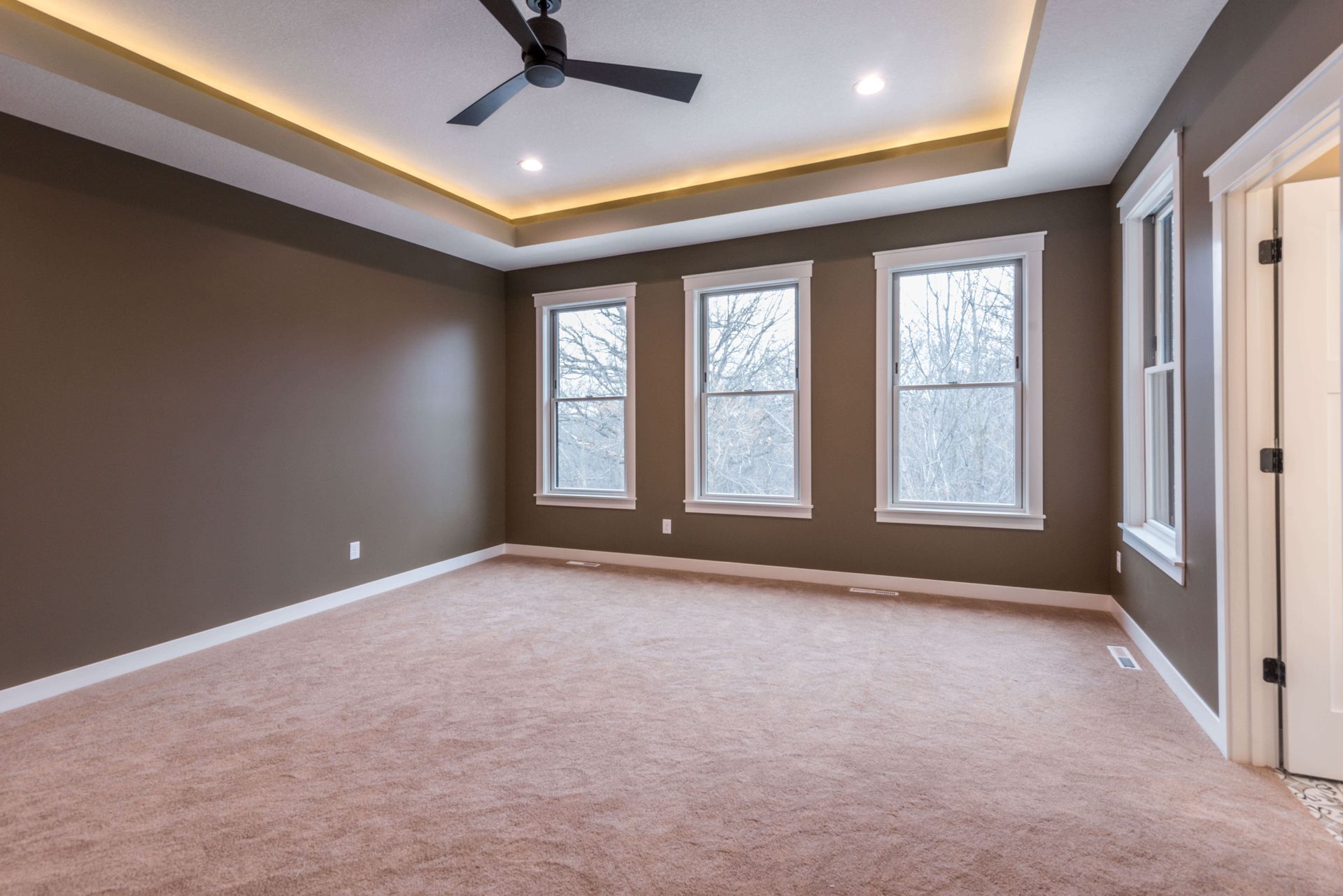 Empty bedroom with brown walls, three windows, beige carpet, and a recessed ceiling with ambient lighting.