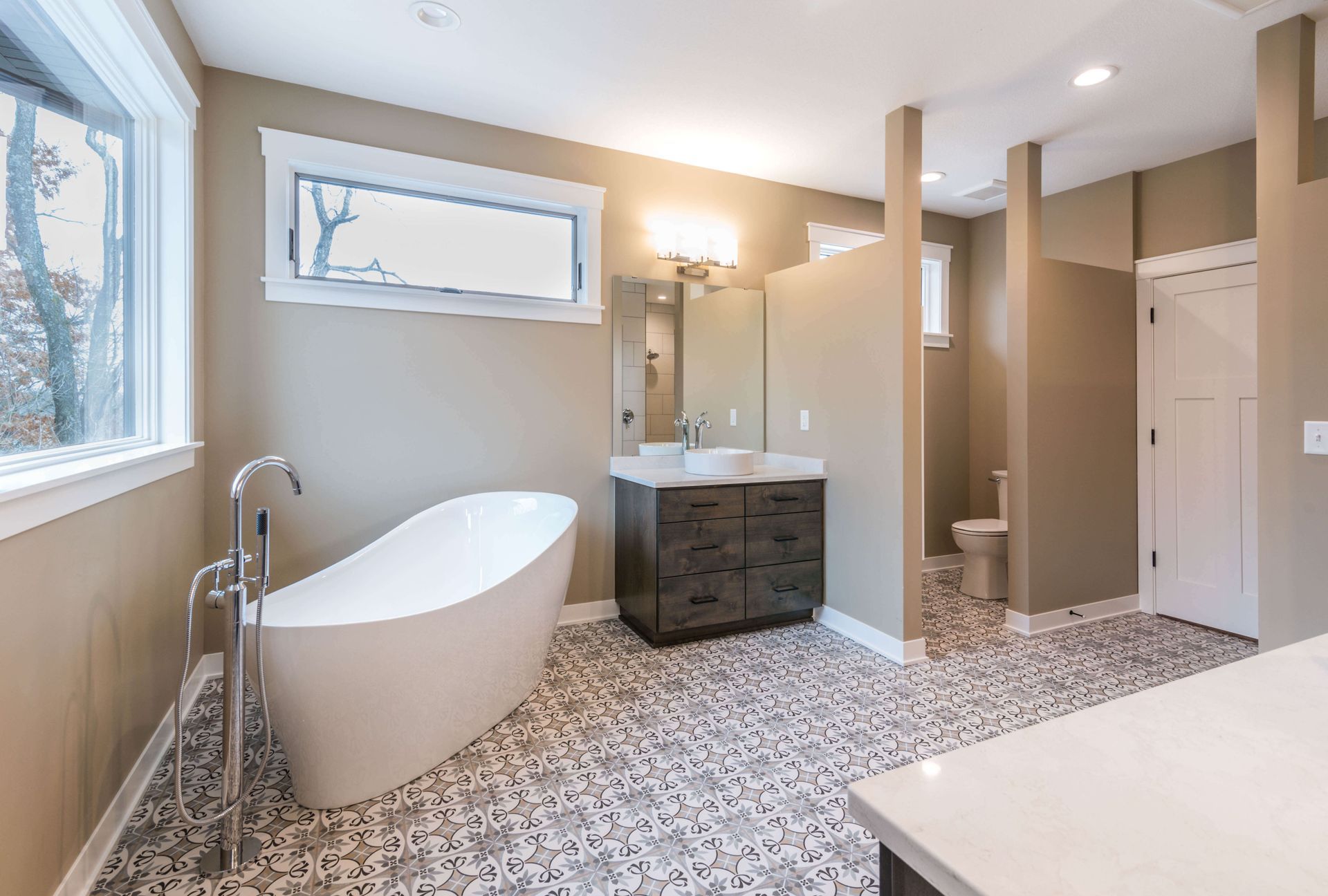 Modern bathroom with a freestanding tub, dark vanity, patterned tile floor, and large windows.