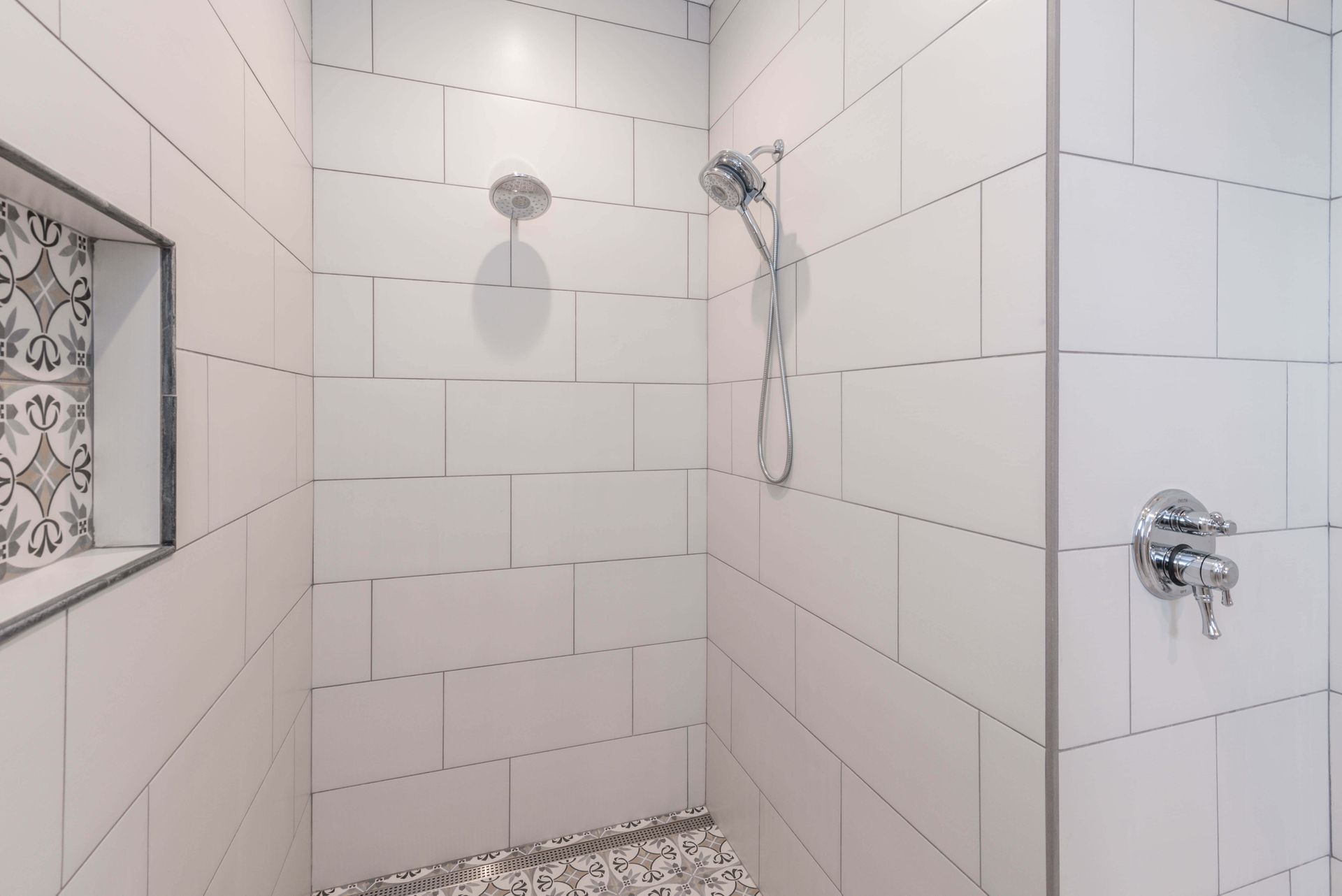 White tiled shower with a decorative inset, silver shower heads, and gray pebble floor.