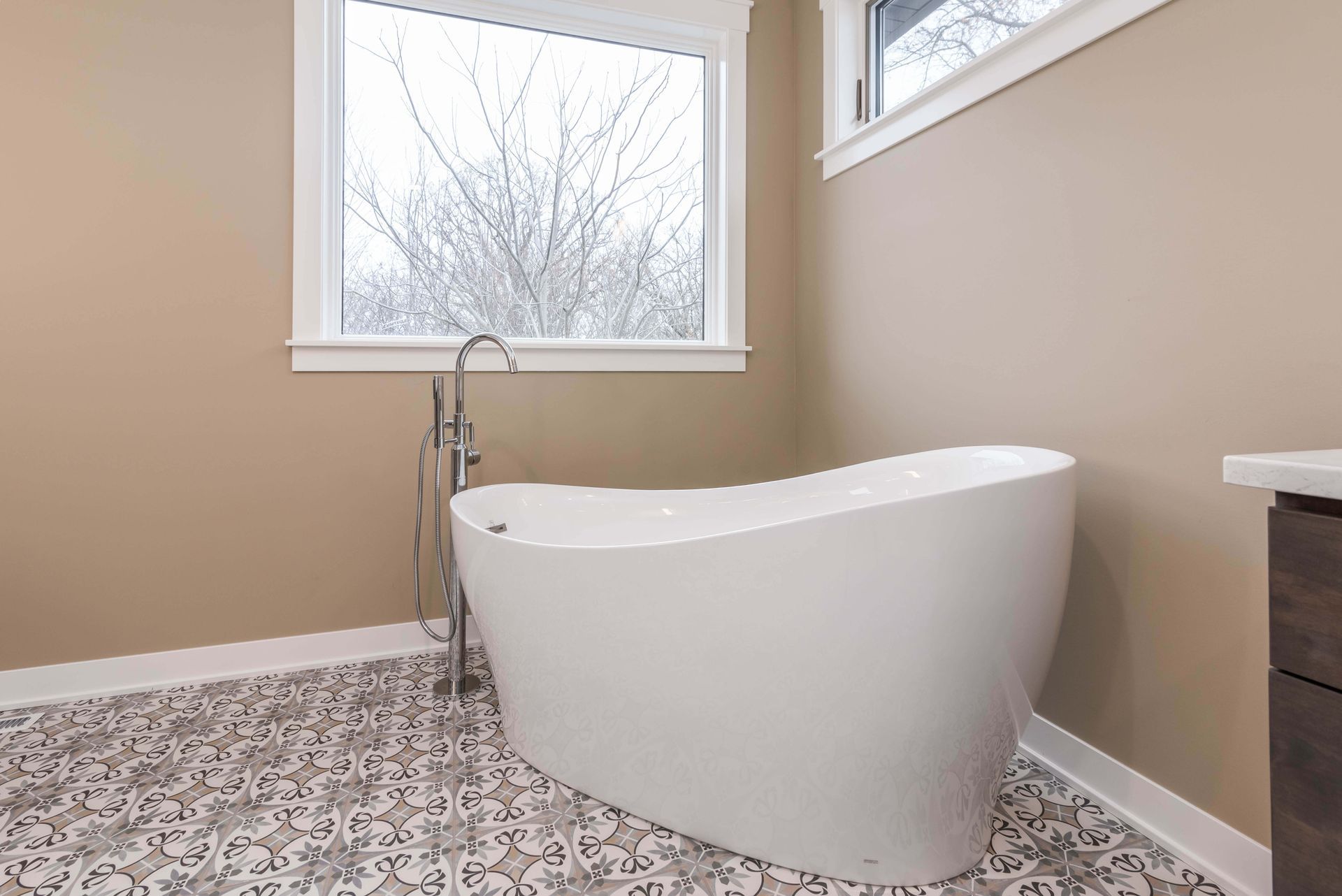 White freestanding bathtub in a bathroom with patterned tile floor, window, and beige walls.