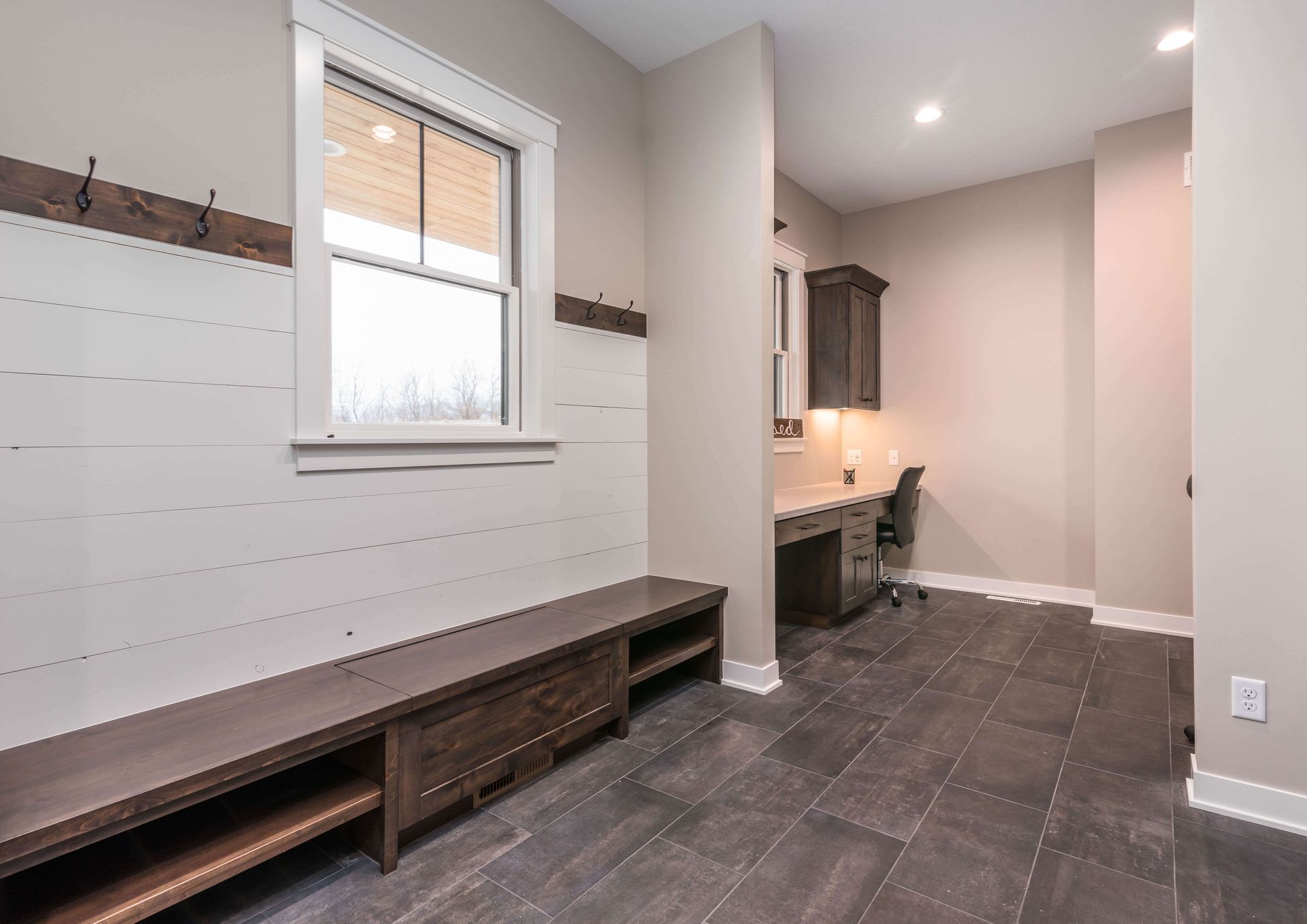 Mudroom with built-in bench, shiplap wall, hooks, window, and desk area with dark wood floors and cabinets.