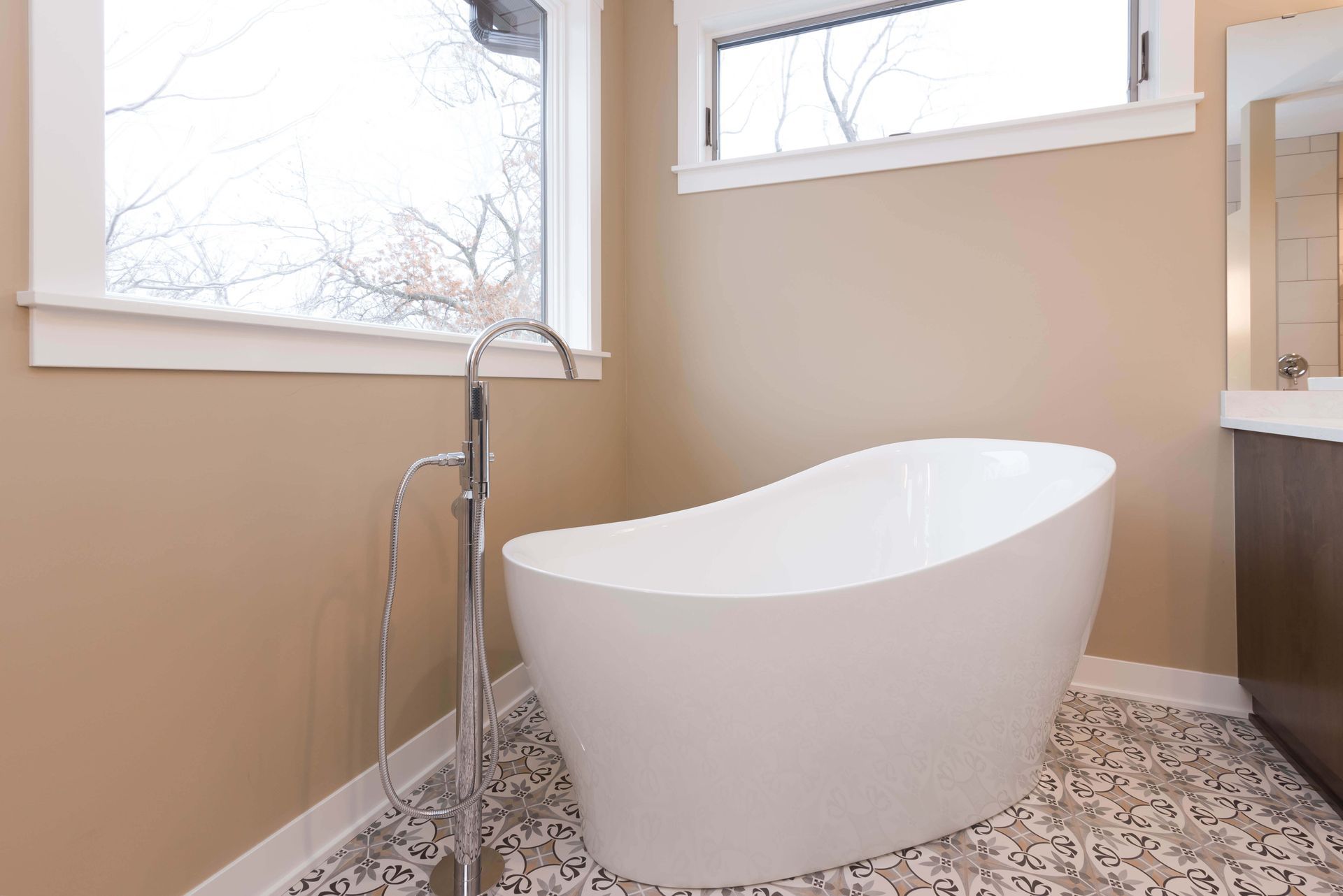 Freestanding white bathtub in a bathroom with a window and a patterned tile floor.