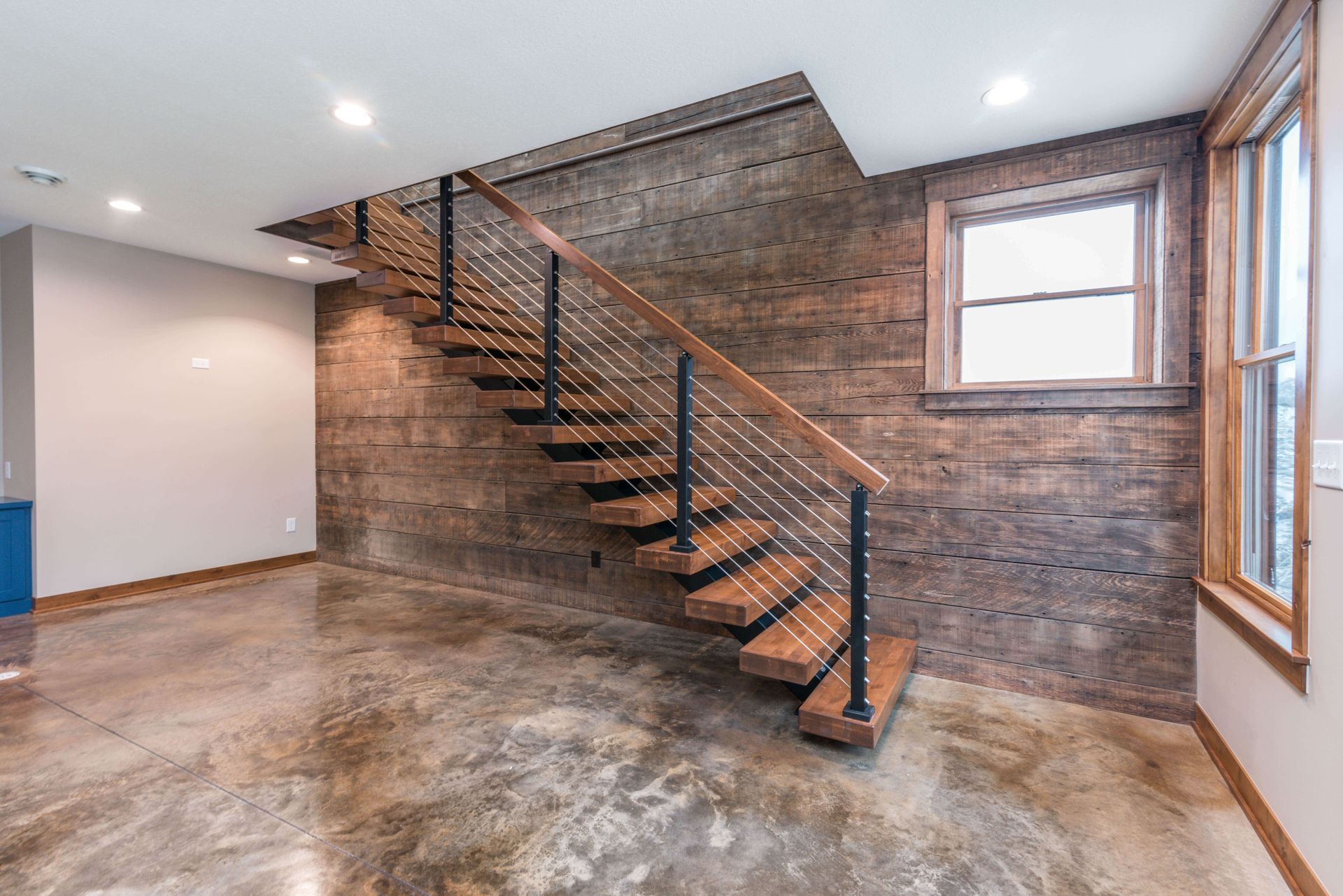 Modern staircase with wooden steps and metal railing against a wood-paneled wall, in a room with a concrete floor.