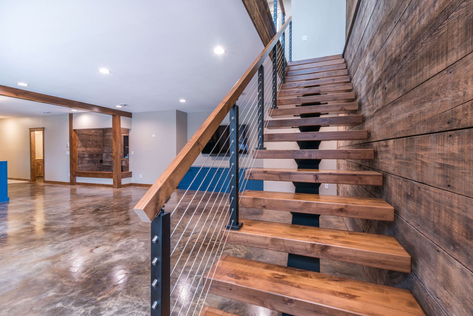 Wooden staircase with metal railing and wood paneled wall. Polished concrete floor and open living area.